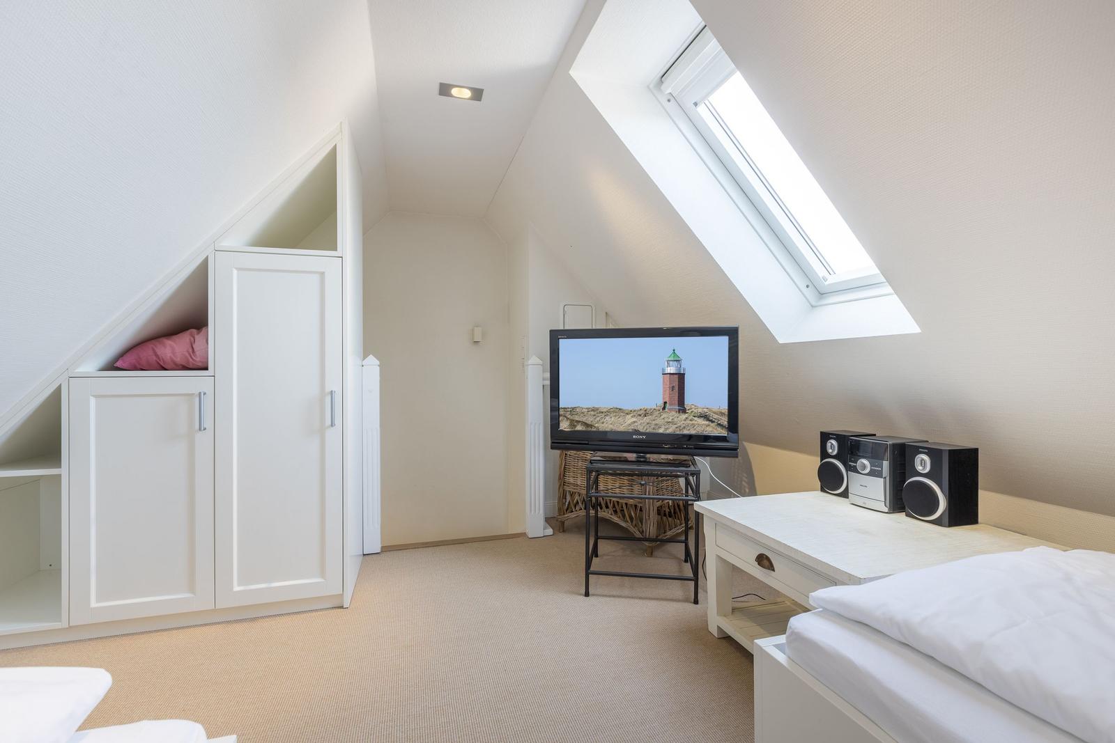 Schlafzimmer mit Dachschräge, TV und Fenster. Weiße Einbaumöbel und Bett.