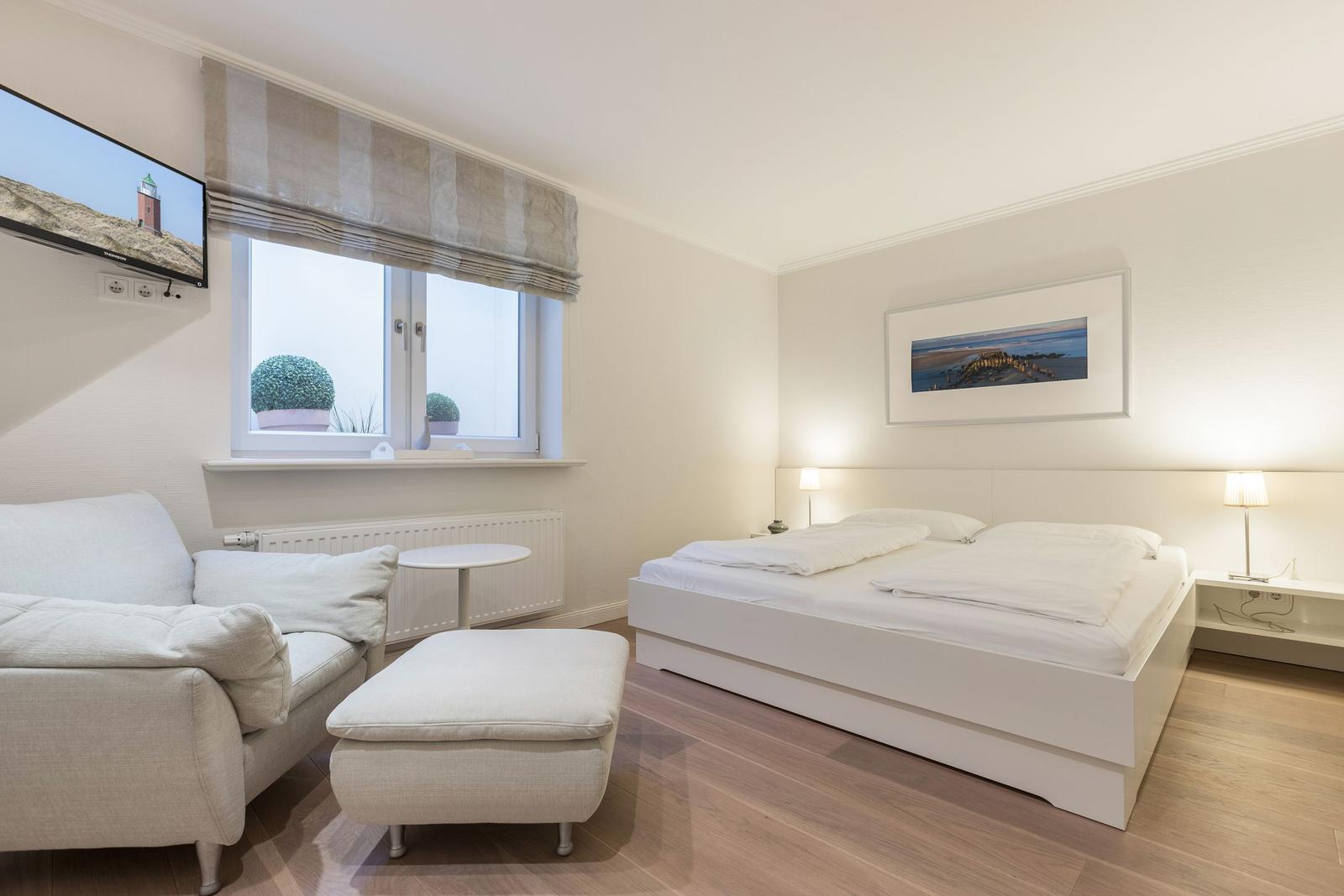Schlafzimmer mit Doppelbett, Sessel und Fernseher an der Wand.