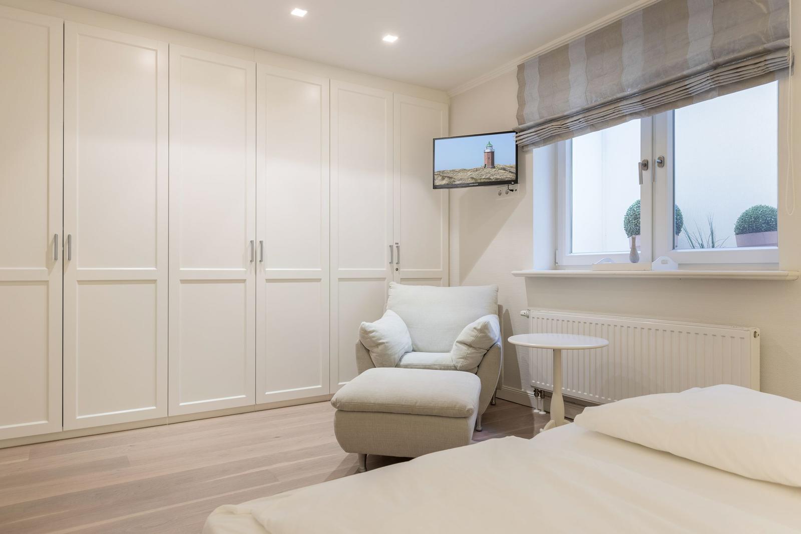 Schlafzimmer mit Bett, Sessel, Kleiderschrank und TV. Fenster mit Rollladen und Pflanzen.