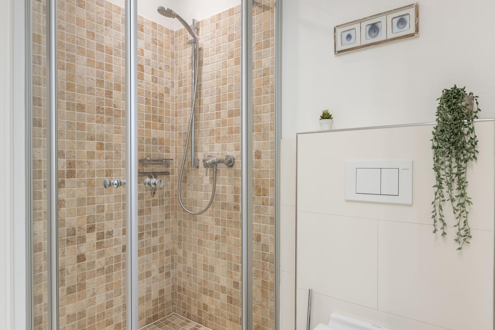 Dusche mit Mosaikfliesen und Duschbrause. Toilette und Pflanzen an der Wand.