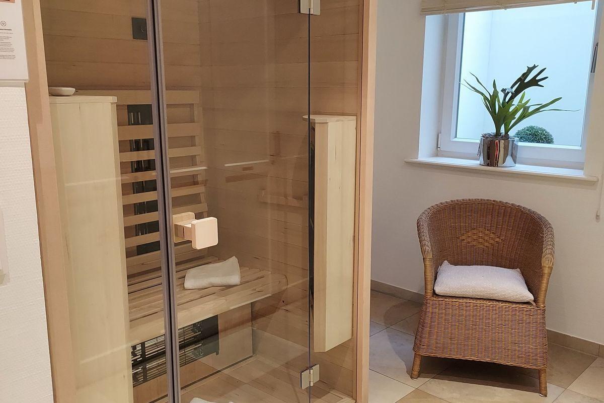 Sauna mit Glaswand und Sitzbank. Nebenher steht ein Rattan-Stuhl mit Kissen.