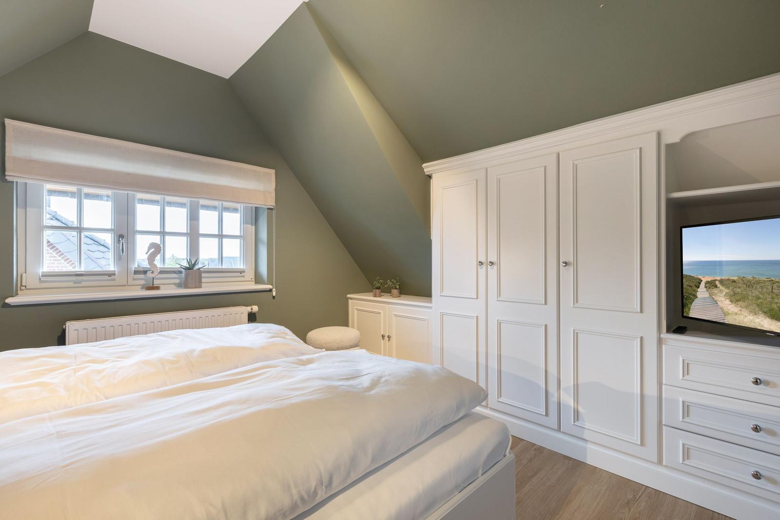 Bedroom with sloped ceiling, white wardrobe, and bed. Window with roller blind and TV.