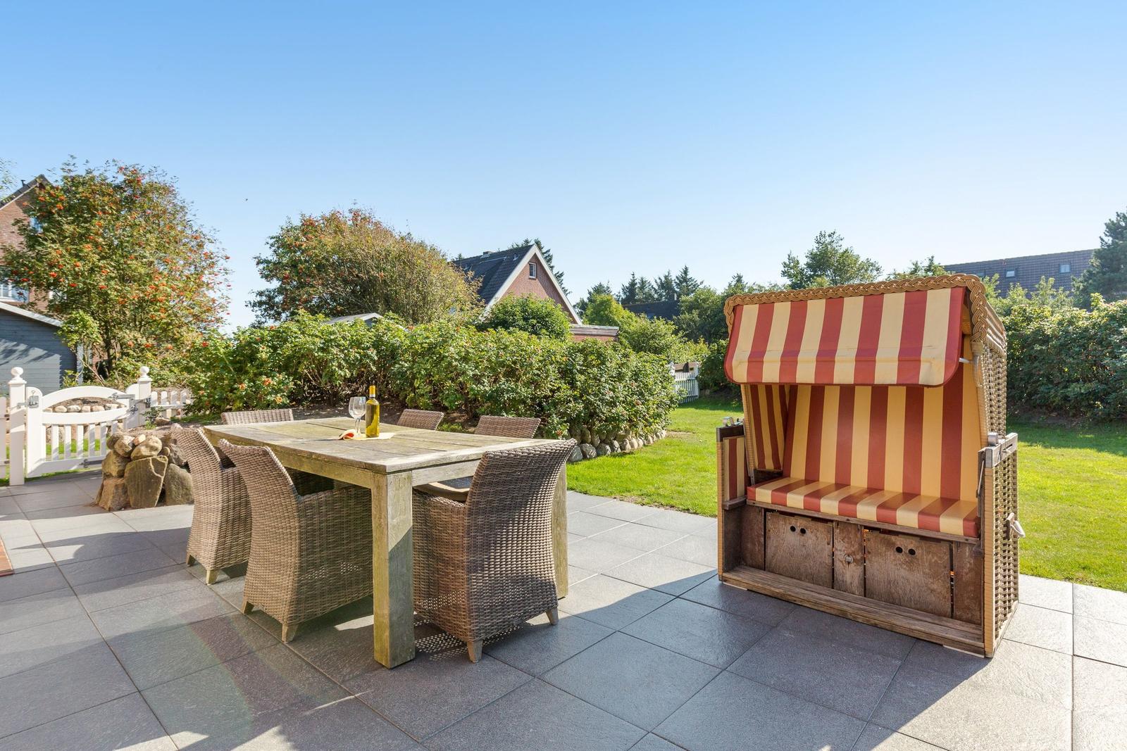 Terrasse mit Esstisch, Stühlen und Strandkorb. Grünflächen und Häuser im Hintergrund.