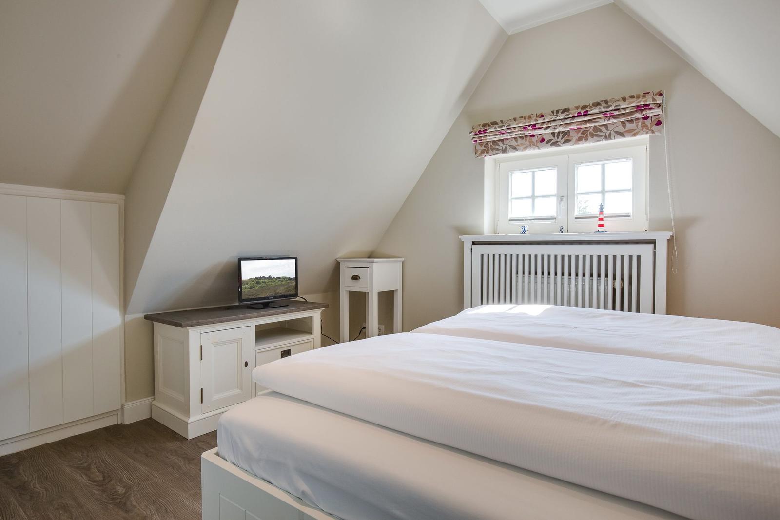 Schlafzimmer mit Dachbodenfenster, weißem Bett und Fernseher auf Schrank.
