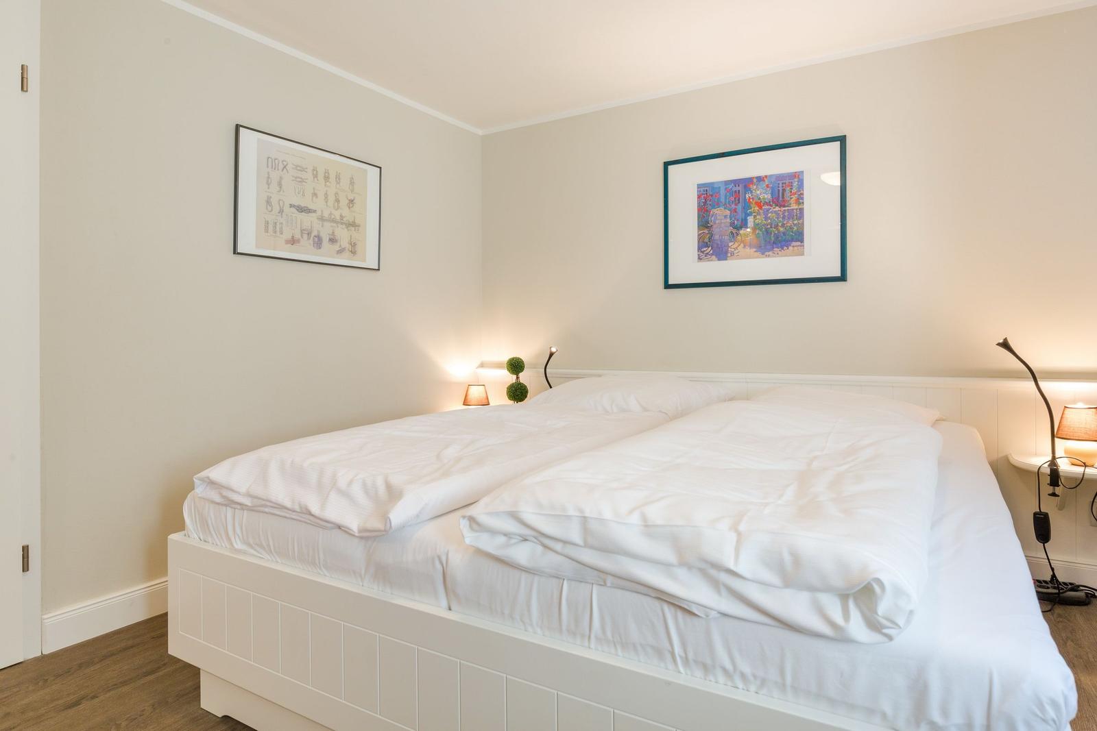 Schlafzimmer mit weißem Bett, zwei Wandbildern und Nachttischlampen.