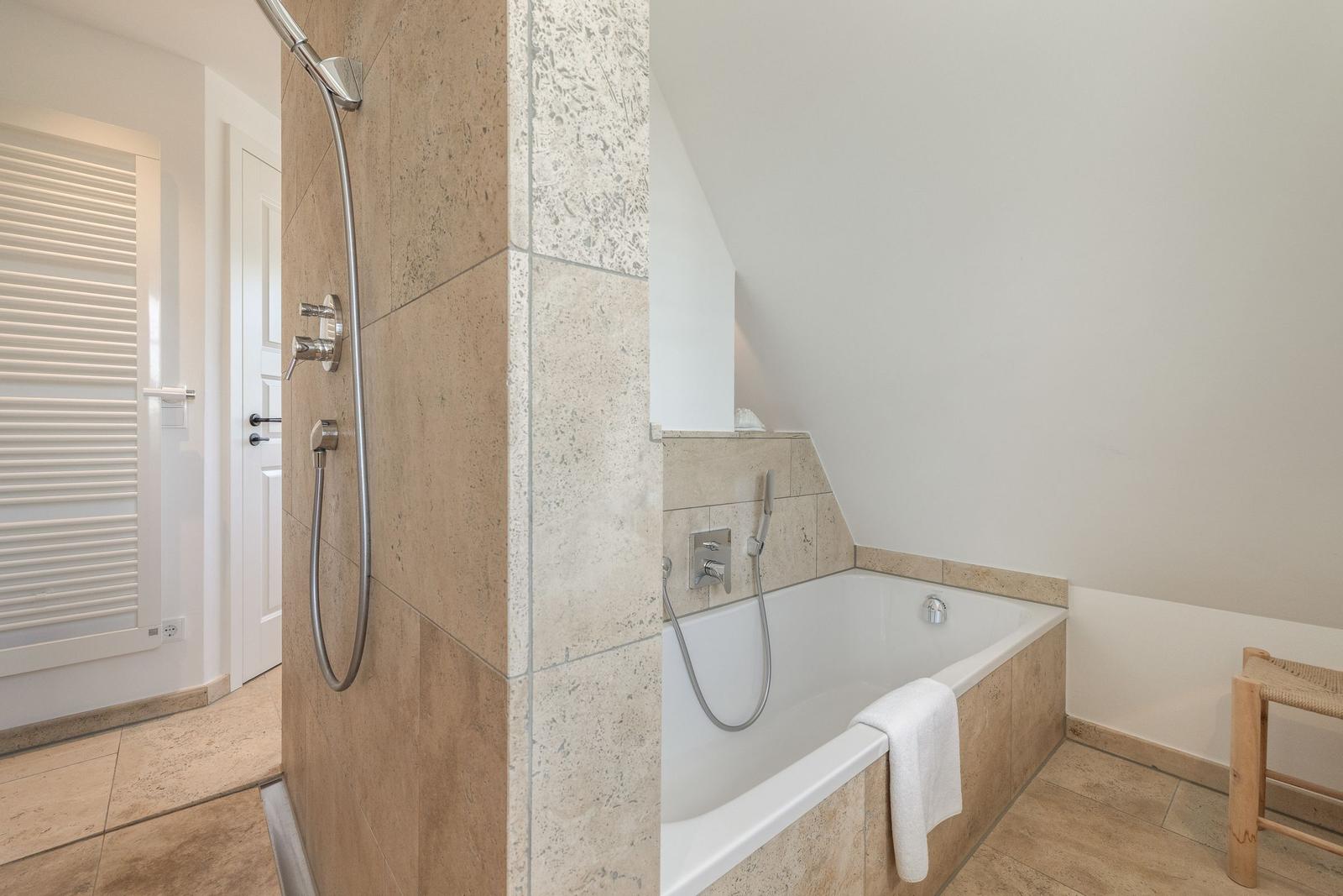 Bath with shower and bathtub, tiled with stone and wooden stool.