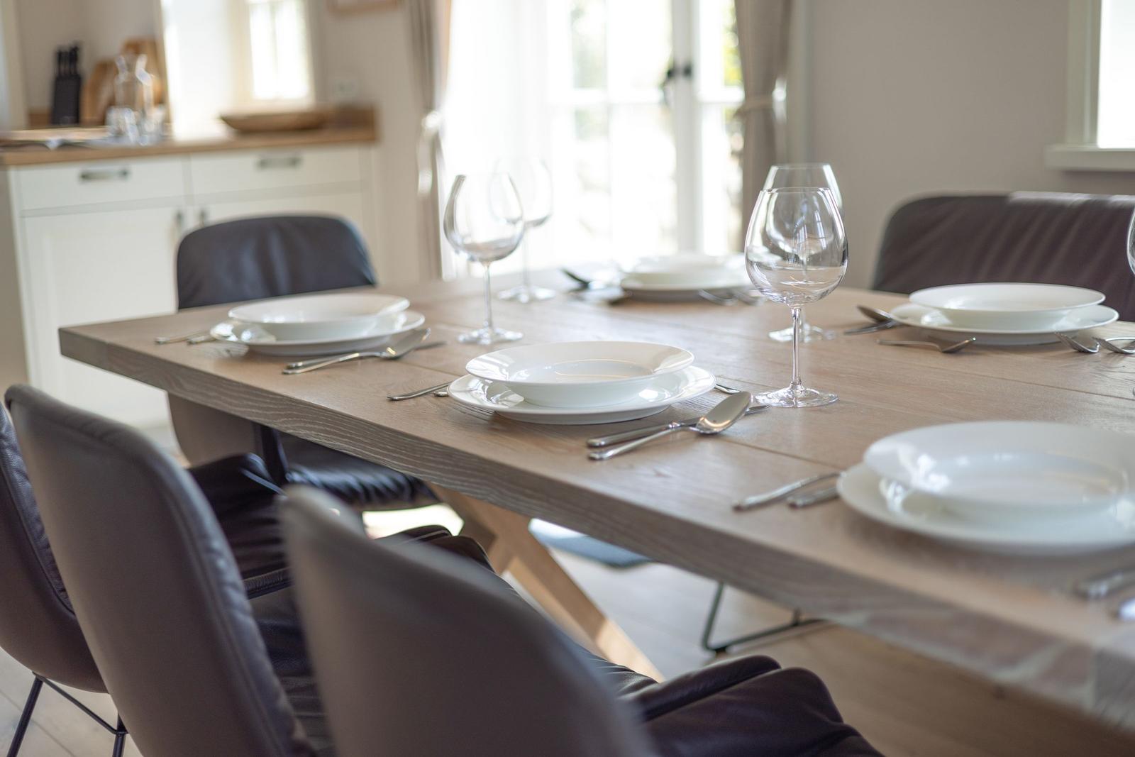 Wooden table with white plates and glasses, dark chairs, kitchen in background