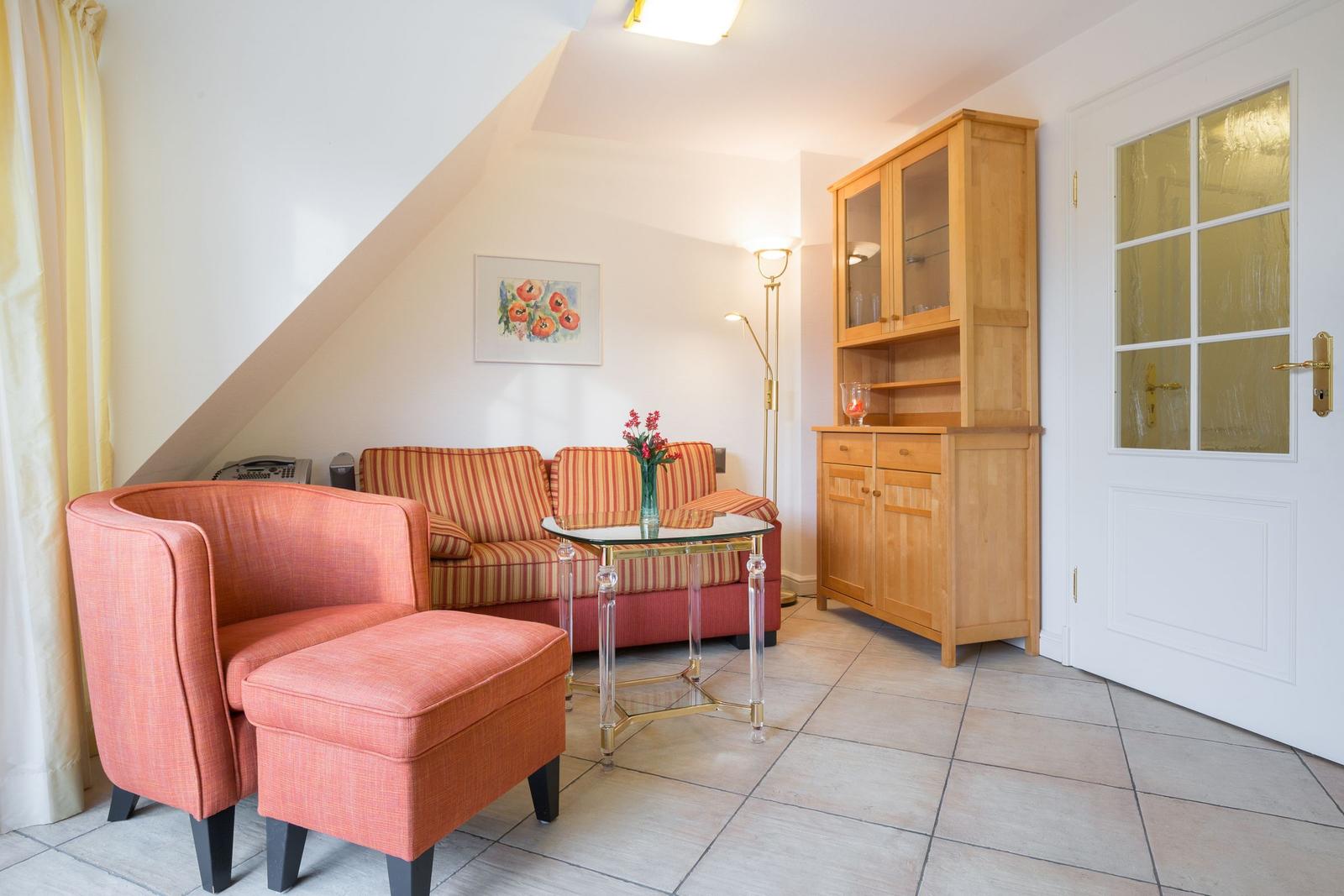 Living area with armchair, sofa, and wooden cabinet. Tile floor and sloped ceiling.