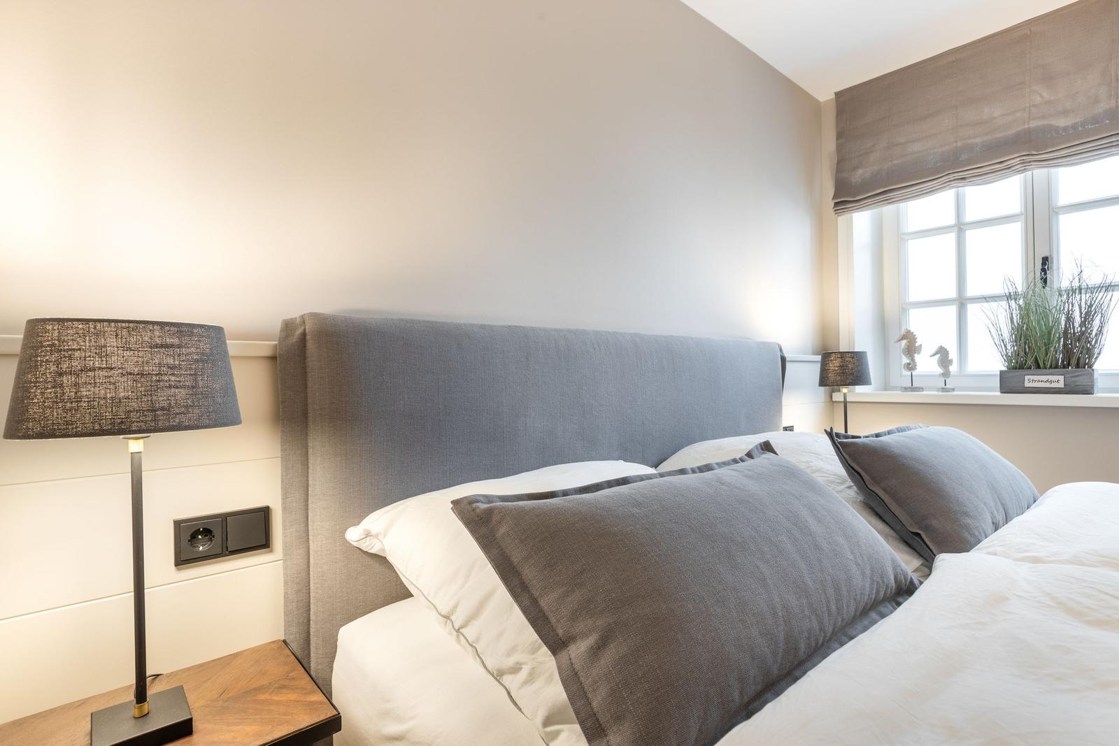 Bedroom with bed, nightstand, and lamp. Window with roller shade and plants.
