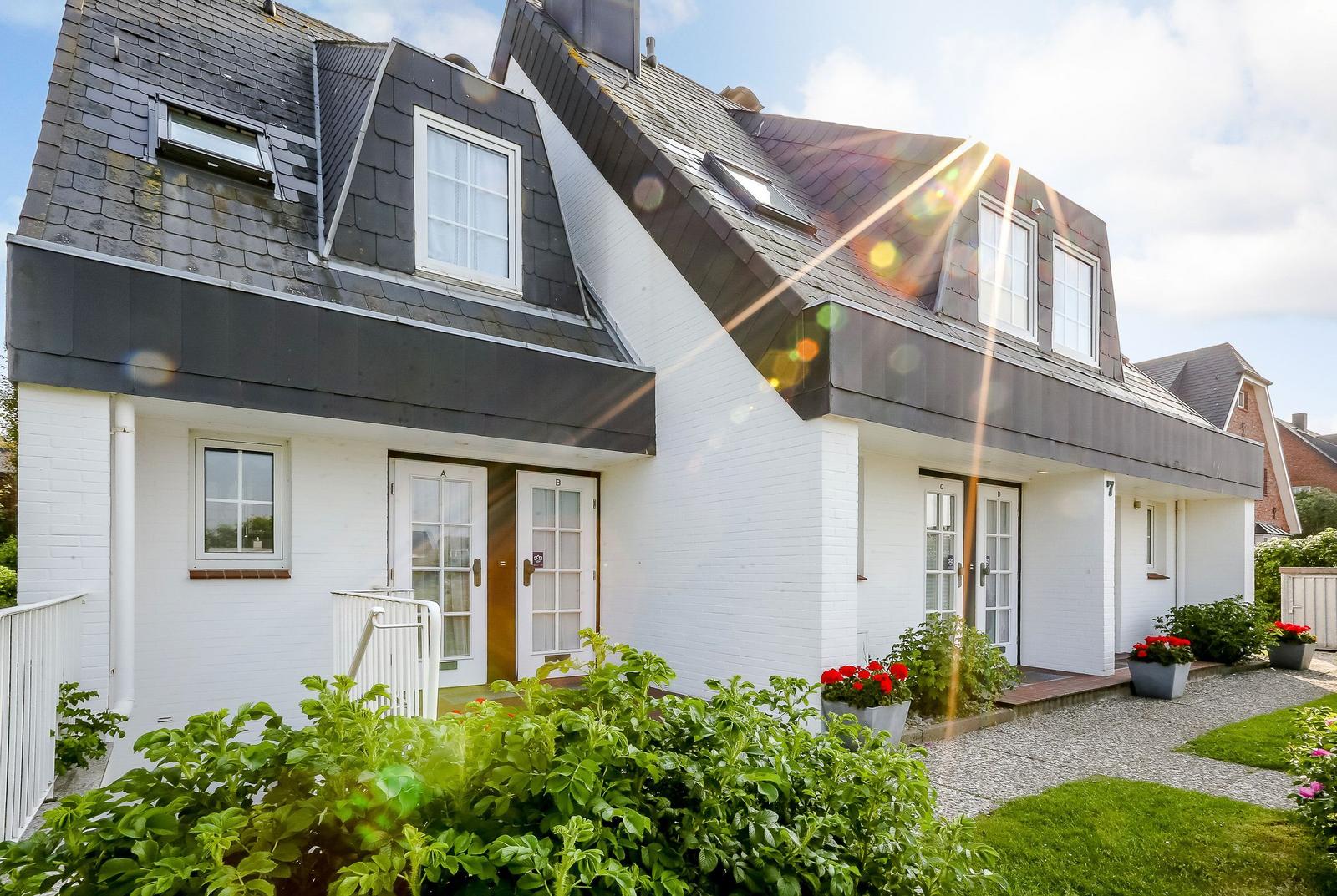 Wohnhaus mit weißen Wänden und dunklem Dach, mehreren Eingängen und Blumenbeeten.