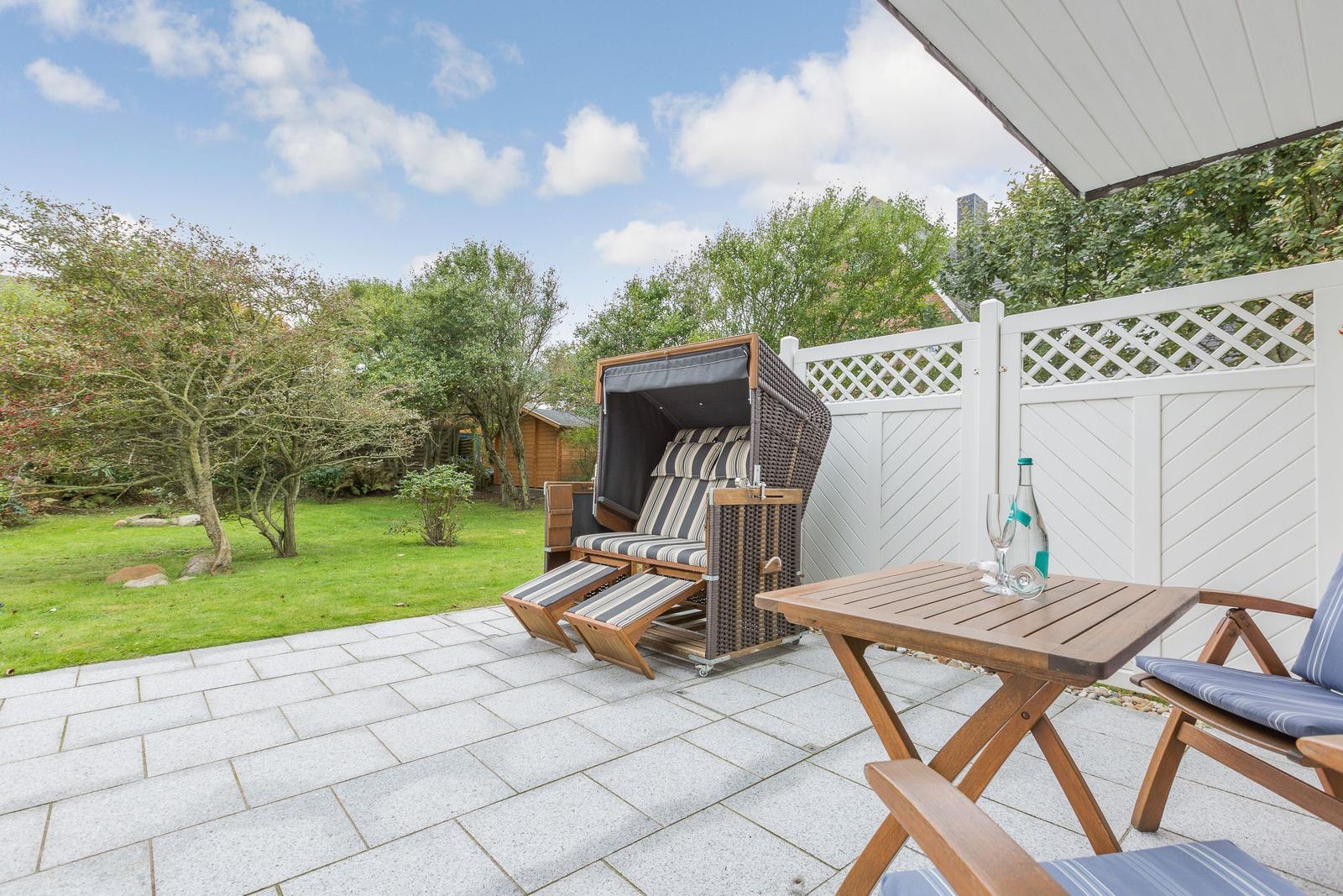 Terrasse mit Liegestuhl, Tisch und Stühlen. Hinterhof mit Grünfläche und weißer Hecke.