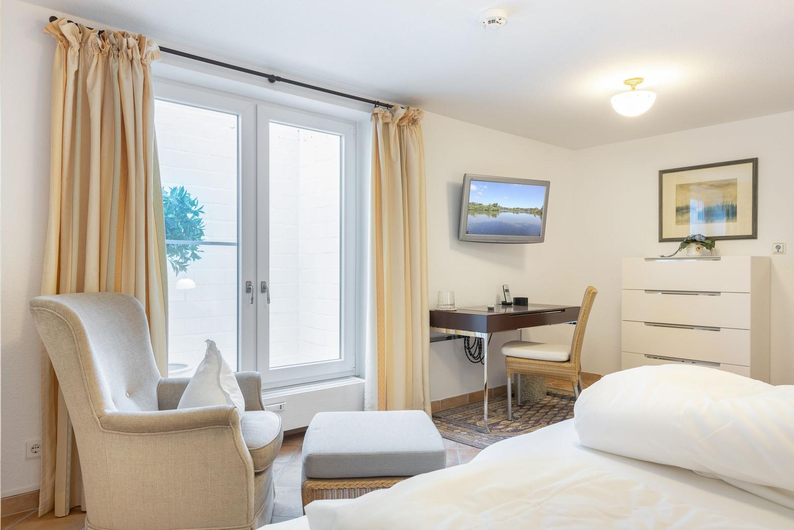 Schlafzimmer mit Bett, Sessel, Schreibtisch und TV. Fenster mit Vorhängen.