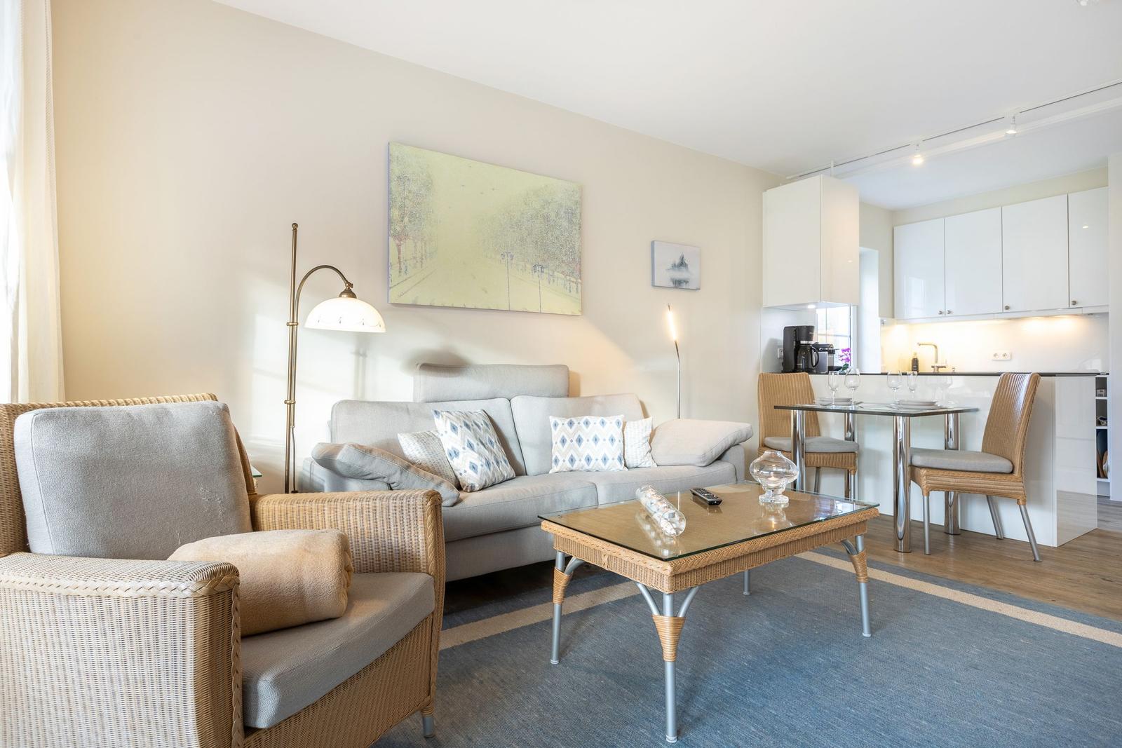 Wohnzimmer mit Sofa, Sessel und Esstisch. Küche mit weißen Schränken.
