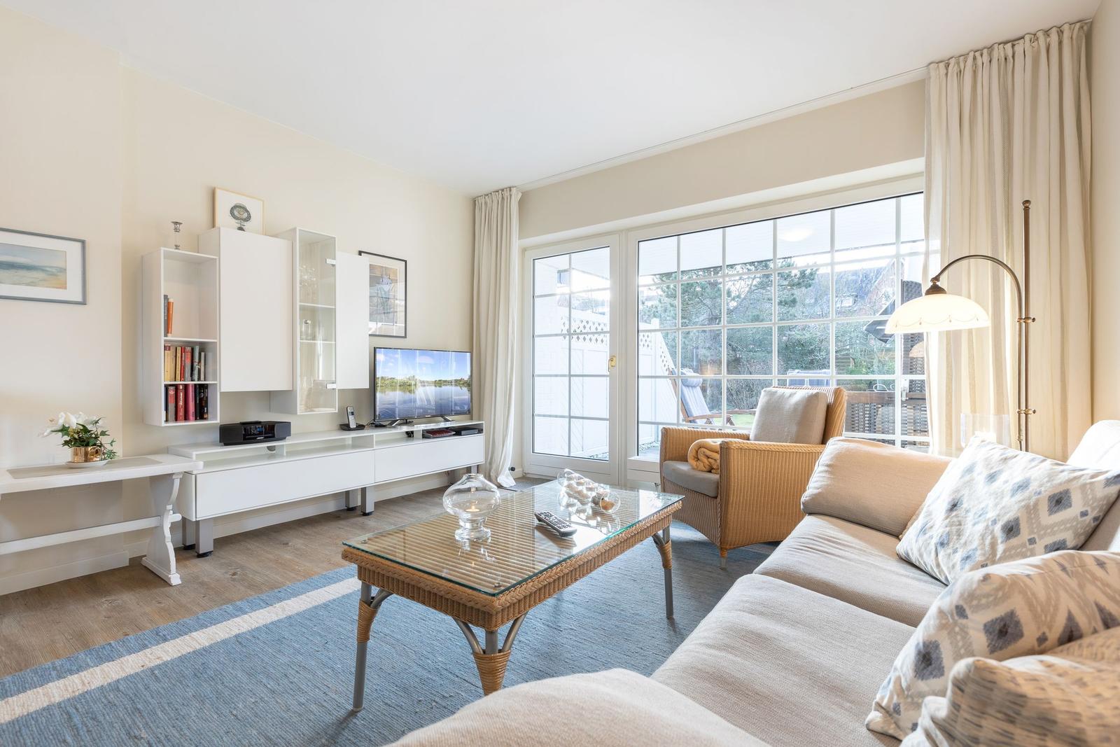 Wohnzimmer mit Sofa, Couchtisch und TV. Große Fenster mit Blick auf Garten.