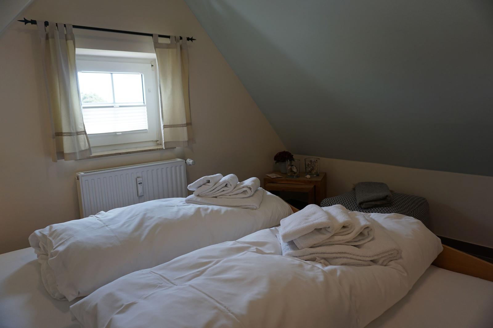 Two beds with white bedding and towels under a sloped ceiling window.
