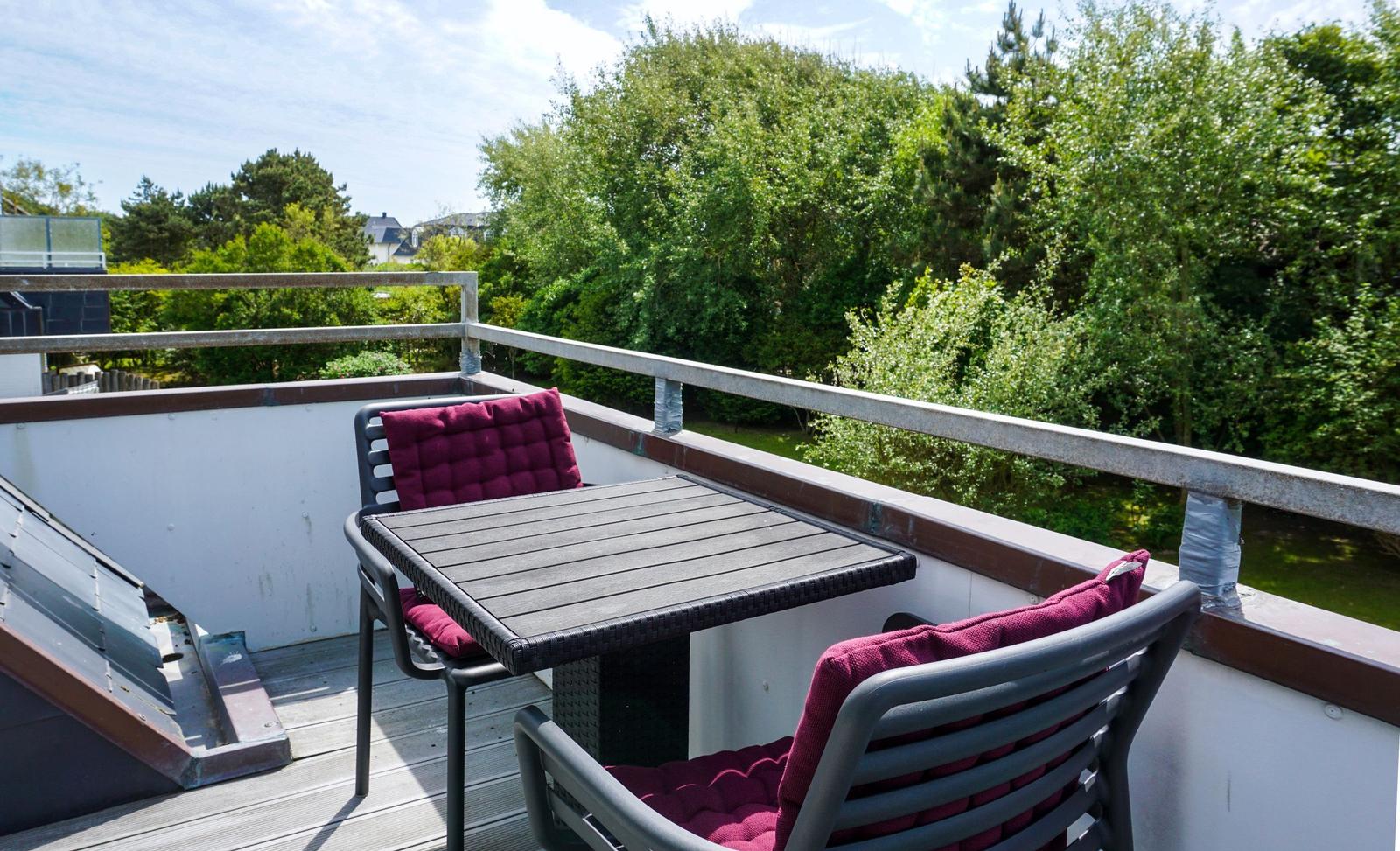 Terrace with table and two chairs with cushions. View of green trees and a river.