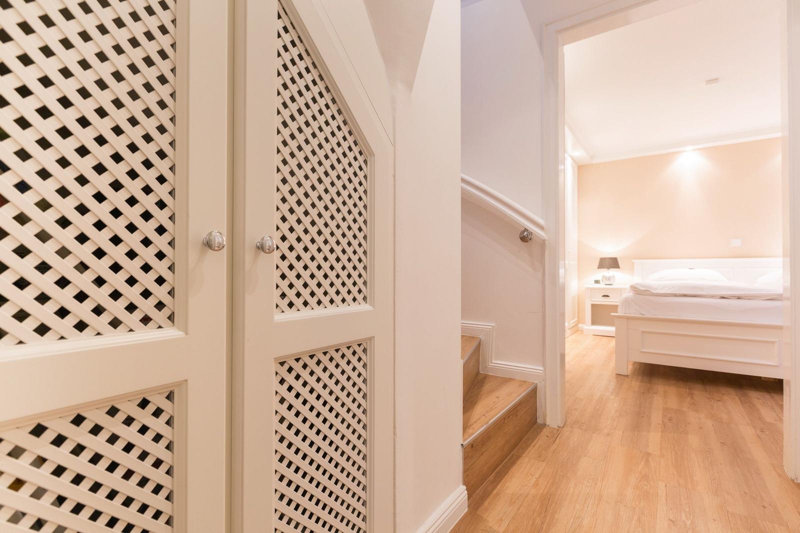 Hallway with wooden stairs and white doors with lattice panels. Bedroom with bed and nightstand in background.