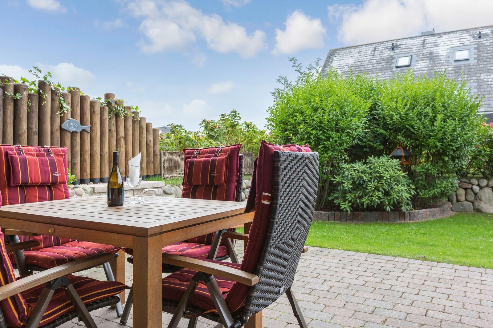 Terrasse mit Holztisch, Stühlen und Weinflasche. Hintergrund: Grünflächen und Holzzaun.