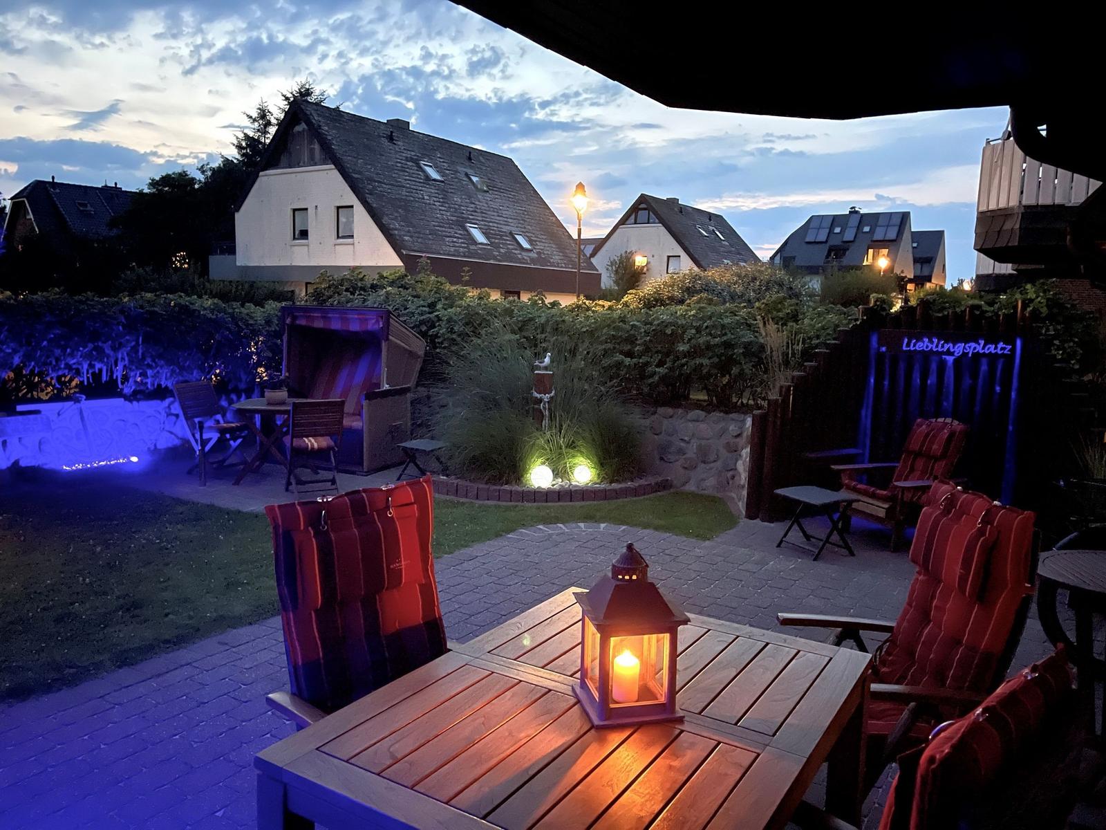 Terrasse mit Tisch, Stühlen und Laterne. Hinterhof mit Garten, Beleuchtung und Haus im Hintergrund.