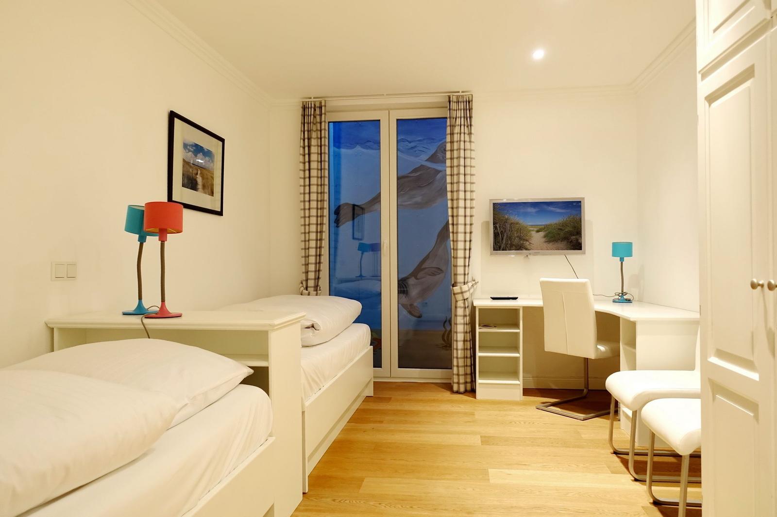 Room with two beds, desk and TV. Windows with curtains.