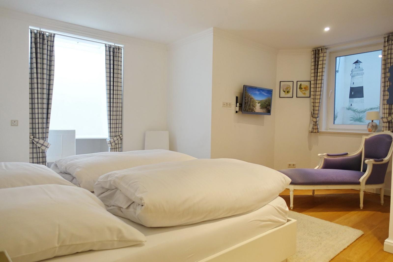 Bedroom with double bed, TV, and chair. Windows with curtains and wall art.