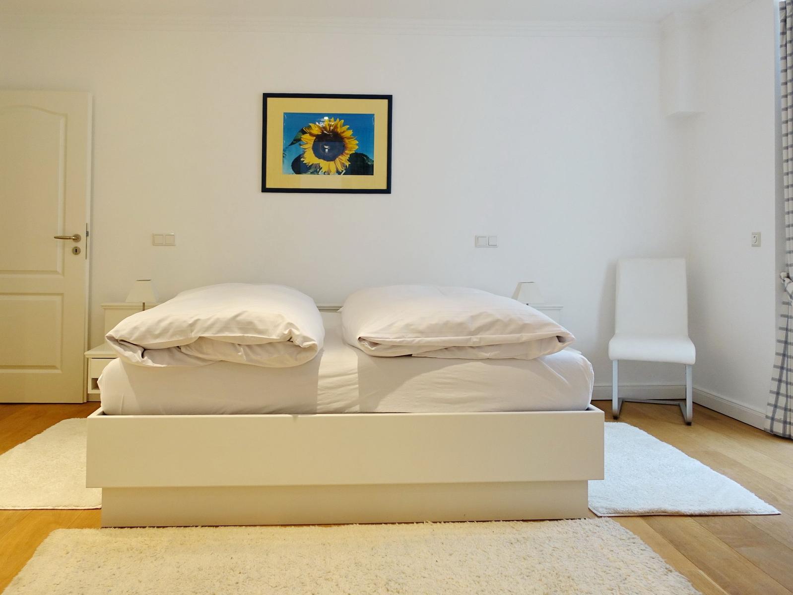 Bedroom with white bed, chair, and wall picture of sunflower.