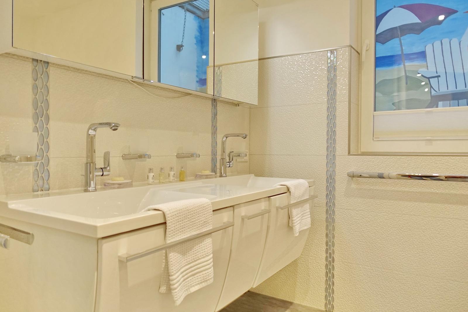 Bathroom with double sink, mirror cabinet, and window with beach picture.