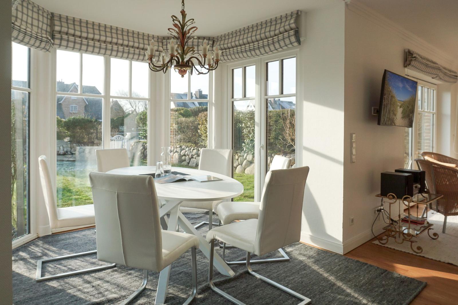 Dining area with round table and white chairs. Large windows overlooking the garden.