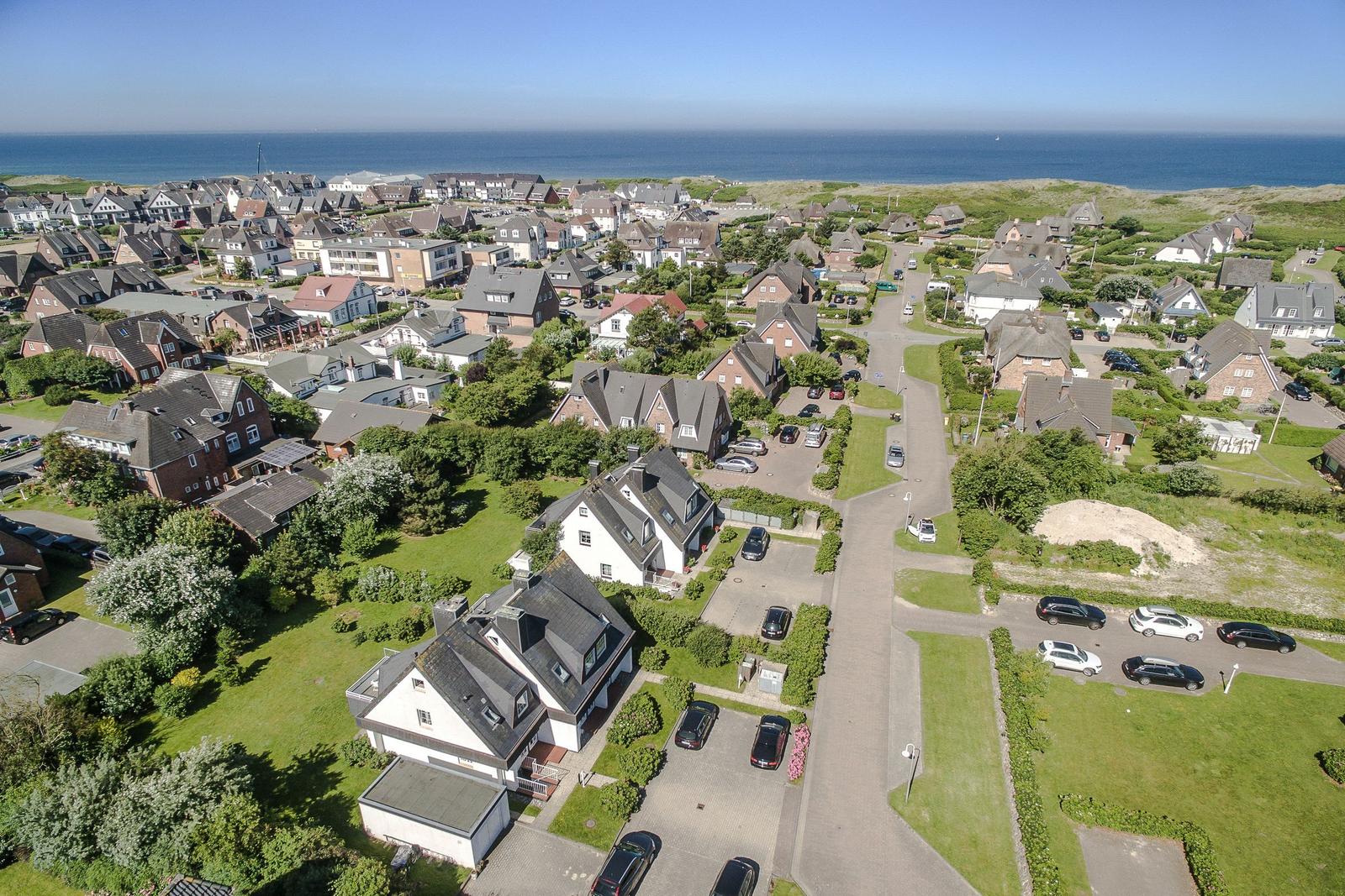 Aerial view of a coastal village with houses, greenery, and the sea in the background.