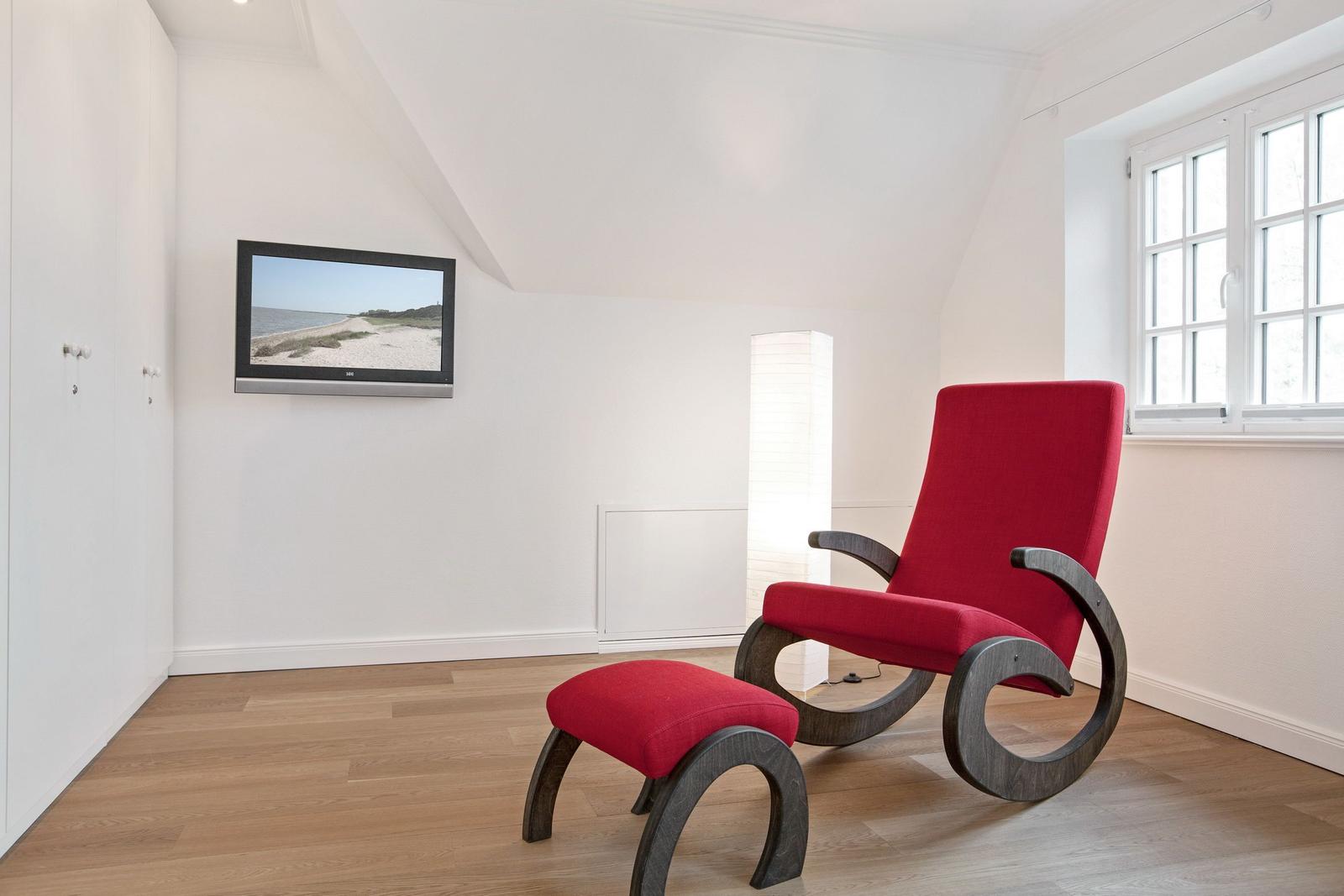 A room with red armchair and footstool, wall-mounted TV, and window.