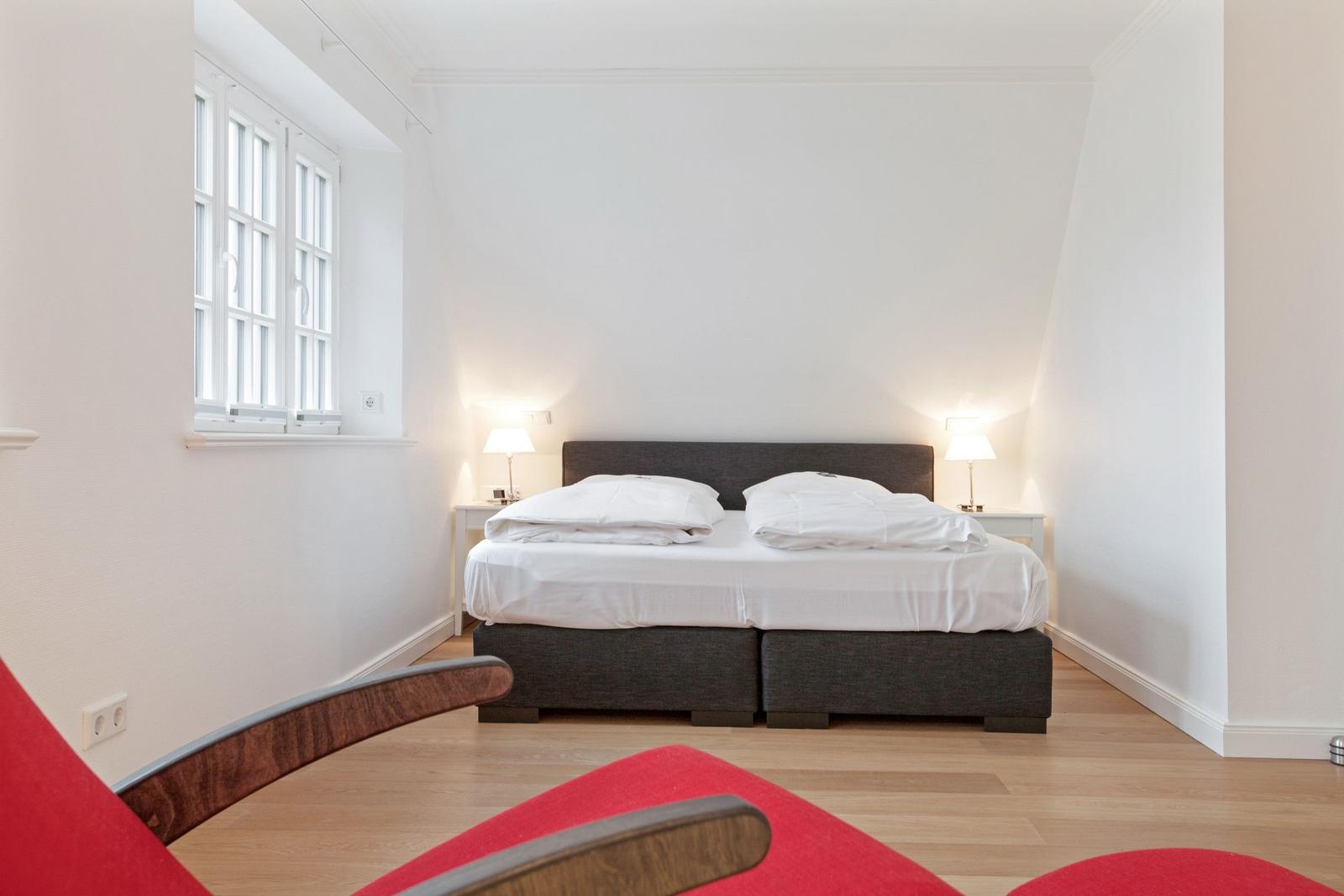 Bedroom with bed, nightstands, and window. Wooden floor and red seat.