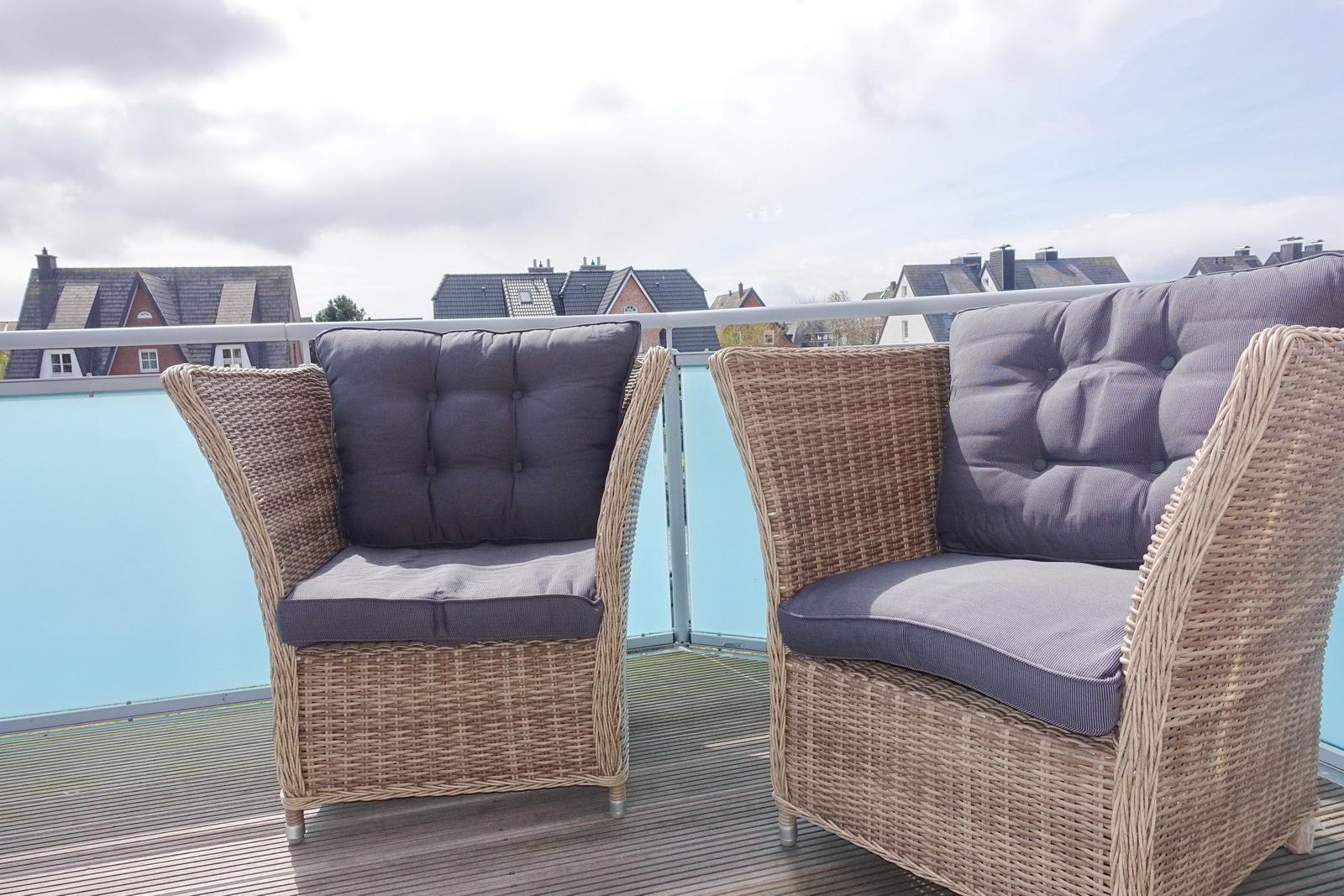 Two wicker chairs with gray cushions on a terrace with a view of houses.