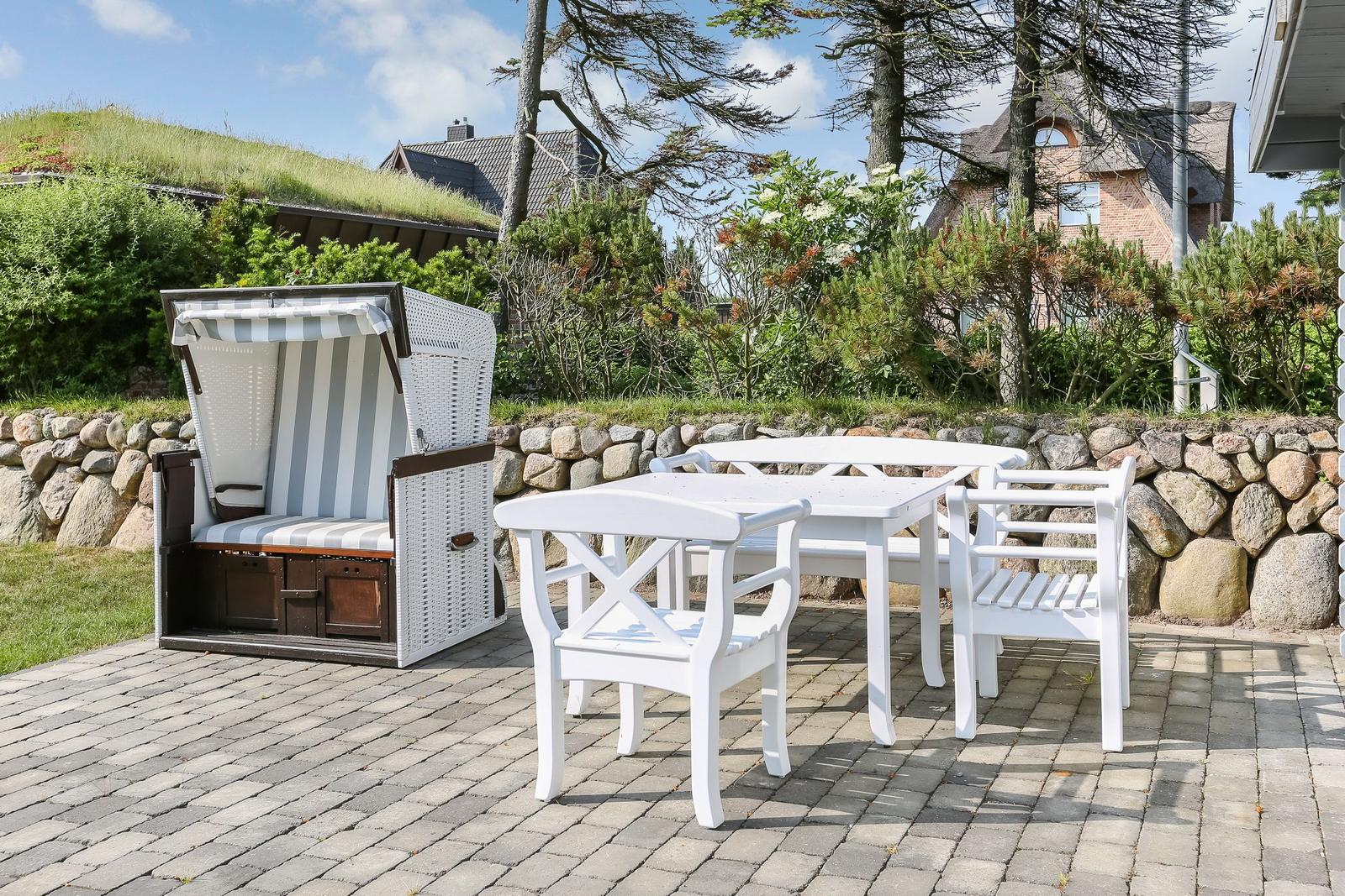 Terrace with white table and chairs and beach chair. Background: stone wall and green areas.