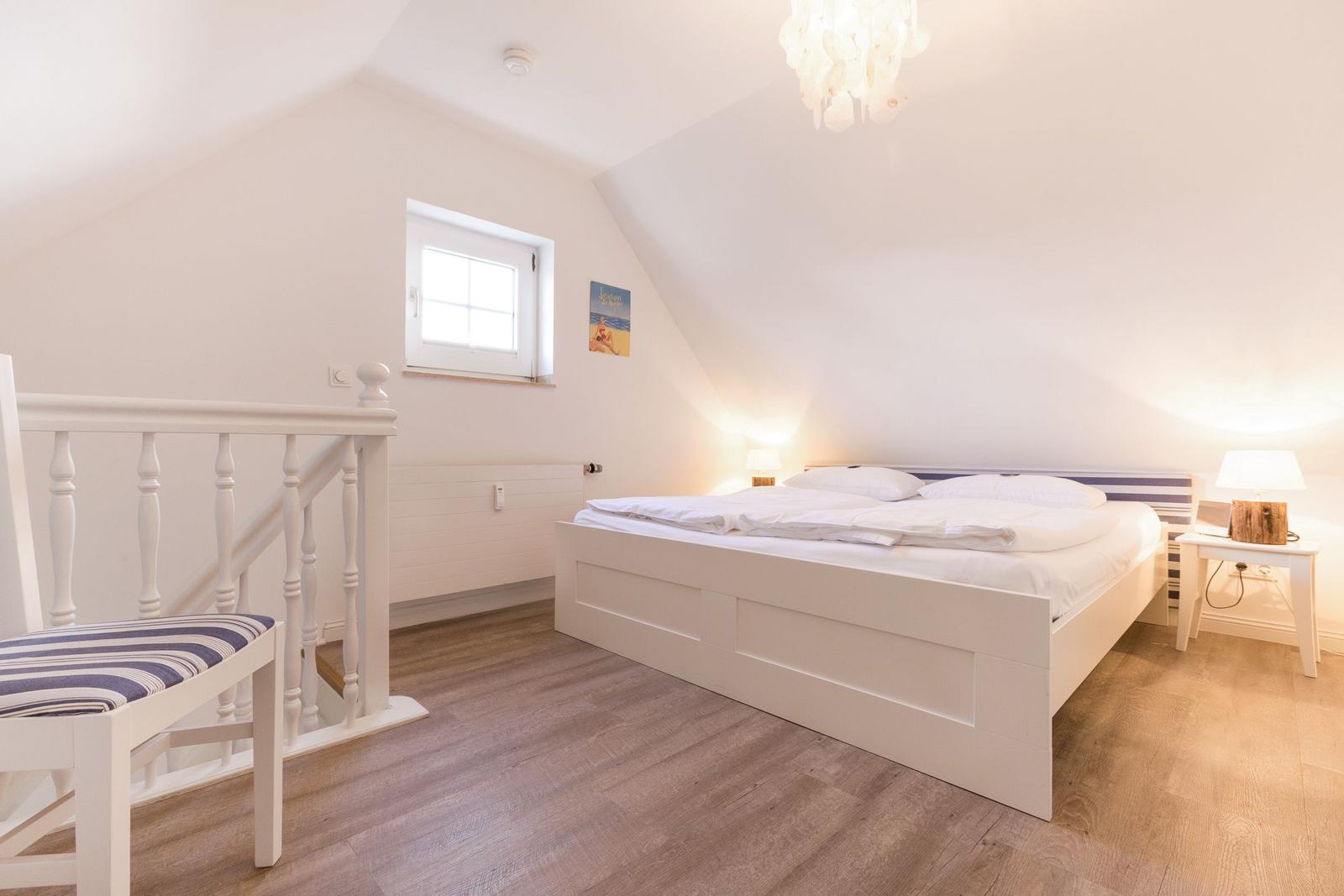 Bedroom with sloped ceiling, white bed, and wooden floor. Window and nightstand visible.