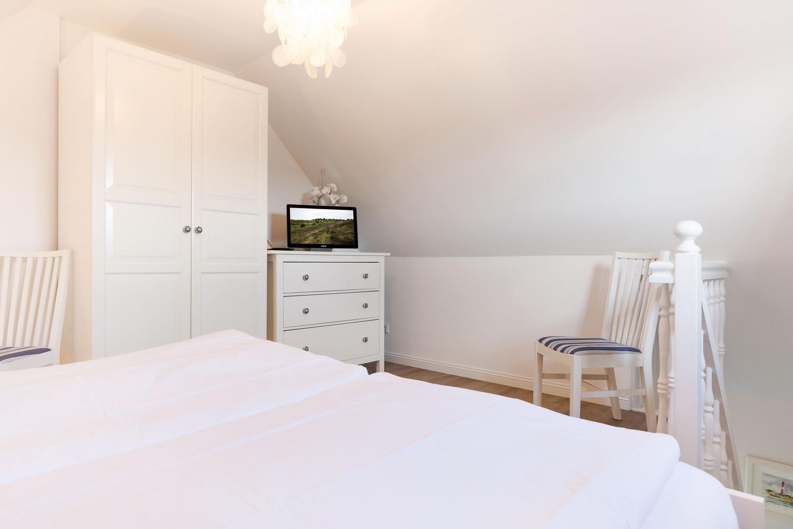 Bedroom with white bed, wardrobe, dresser, and chair under sloped ceiling.