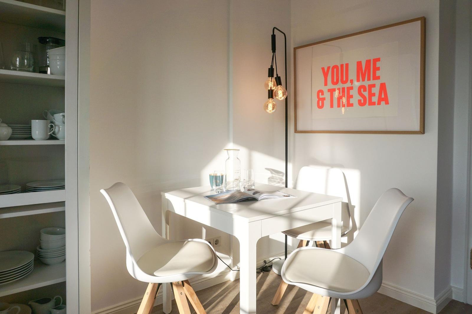 White dining set with chairs and wall art. Dishware in open cabinet.