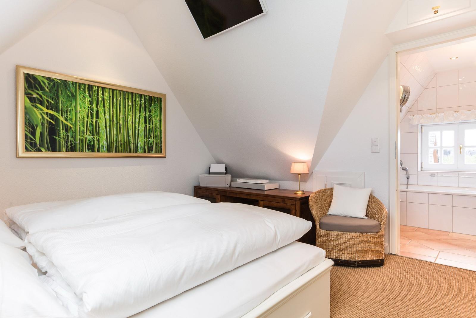 Schlafzimmer mit Dachschrägen, Bett, Wickerstuhl und Bad im Hintergrund.