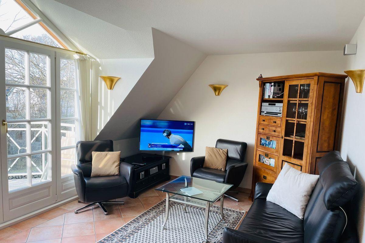 Wohnzimmer mit TV, zwei Sesseln, Sofa und Glasplatte. Fenster zur Terrasse.