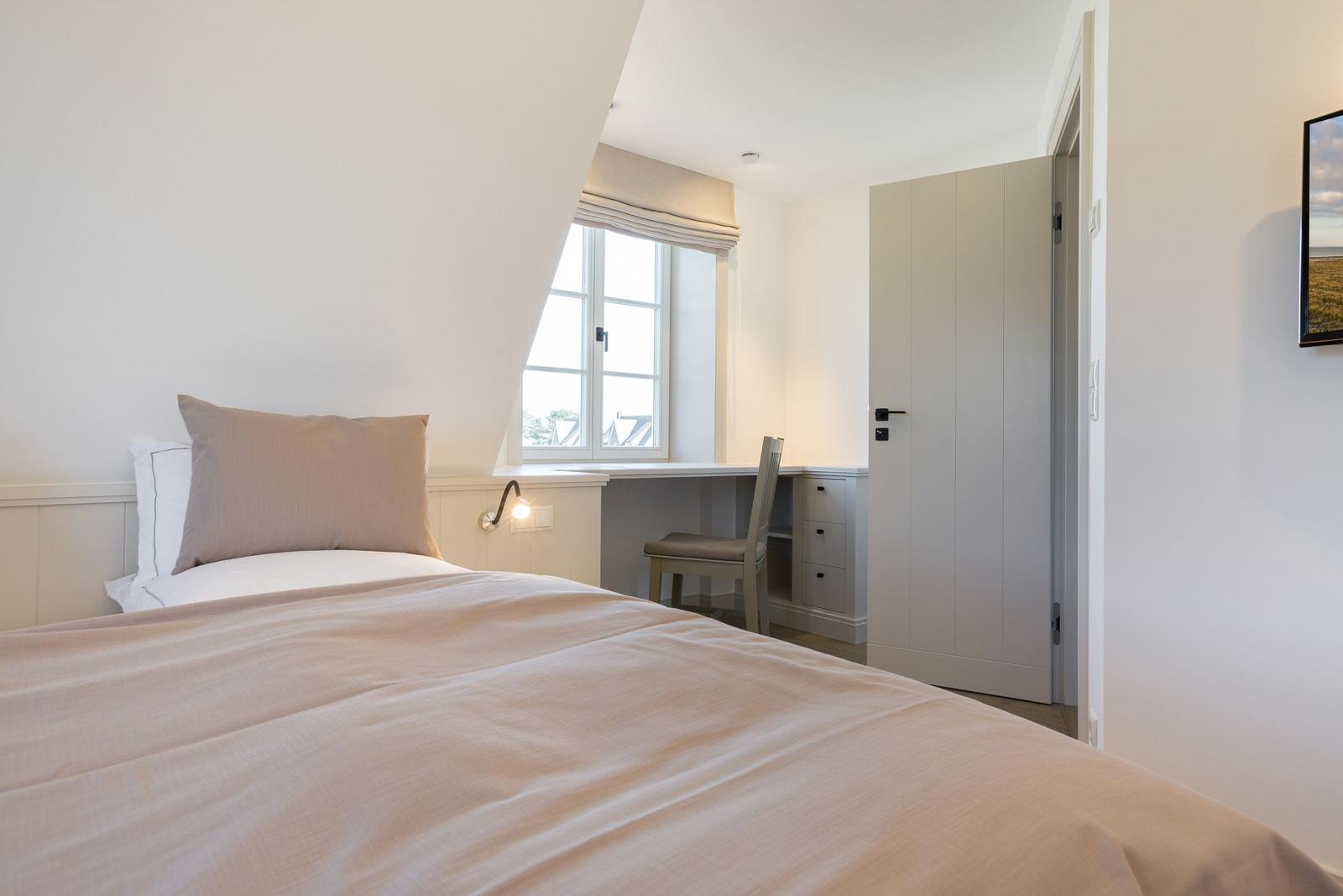 Schlafzimmer mit Bett, Schreibtisch und Fenster. Tür führt zu einem Ankleidezimmer.