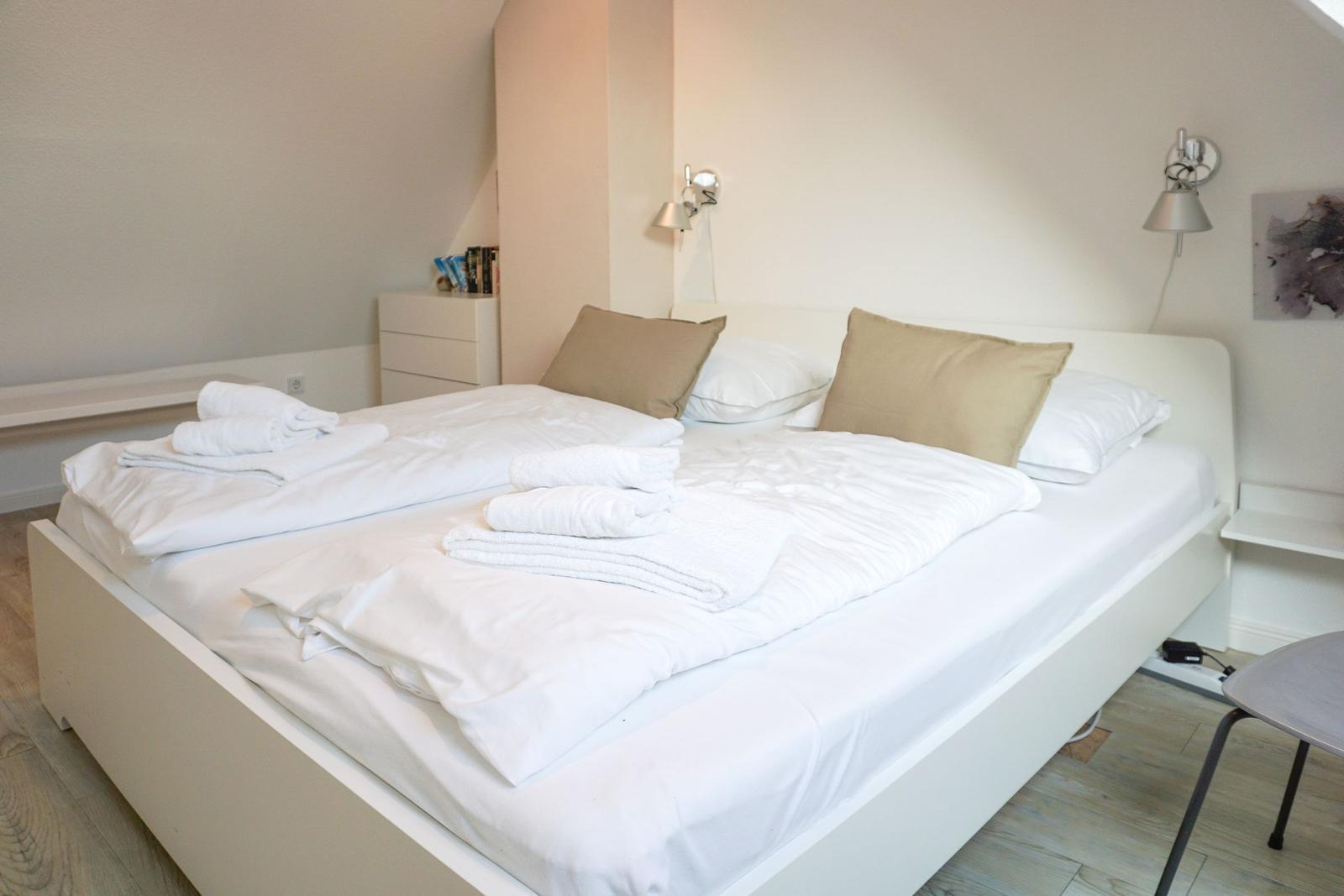 Bedroom with white bed, pillows, and towels. Wall lamps and wooden floor.