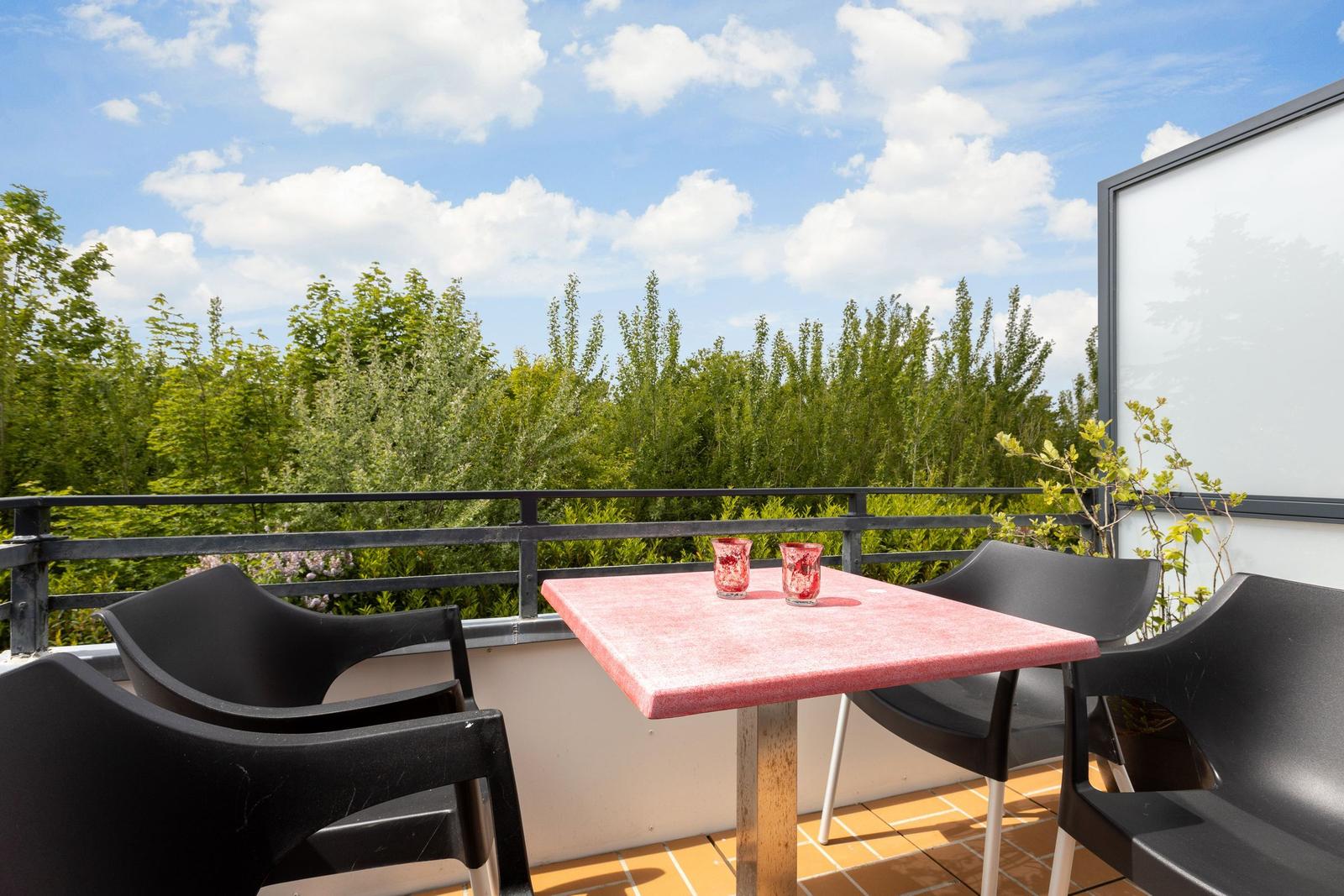 Terrace with table, chairs, and view of green trees under blue sky.