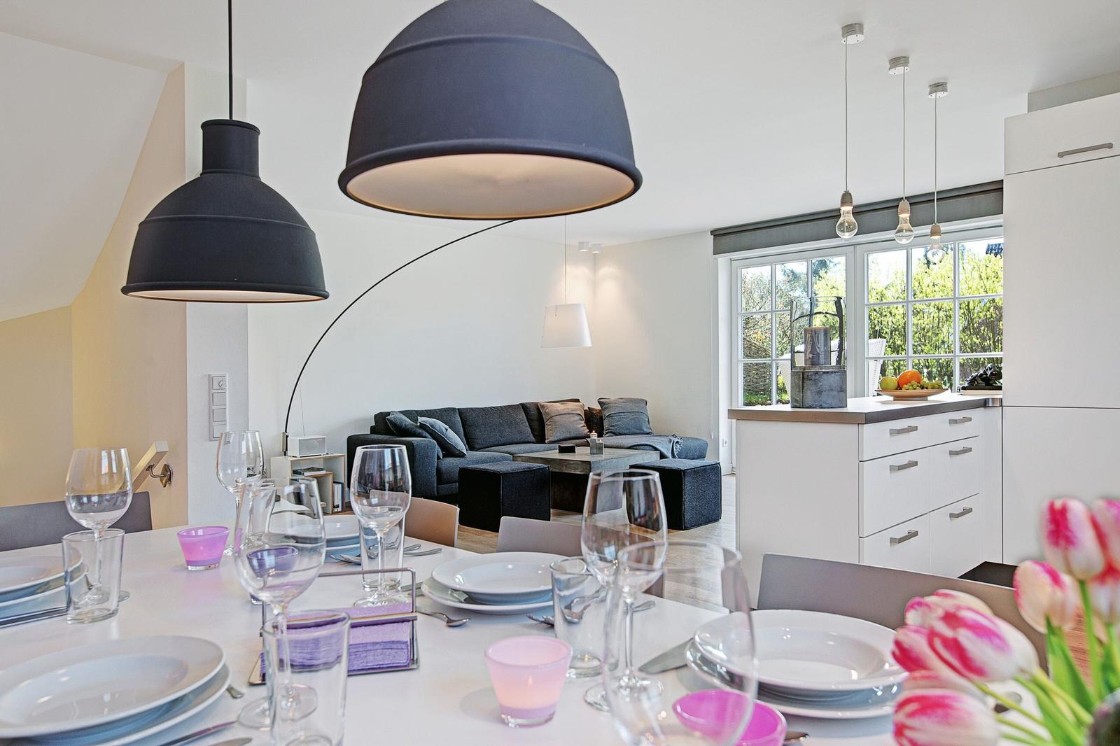 Essbereich mit Tisch, Stühlen und großen Fenstern. Küche mit weißen Schränken. Wohnbereich mit Sofa.