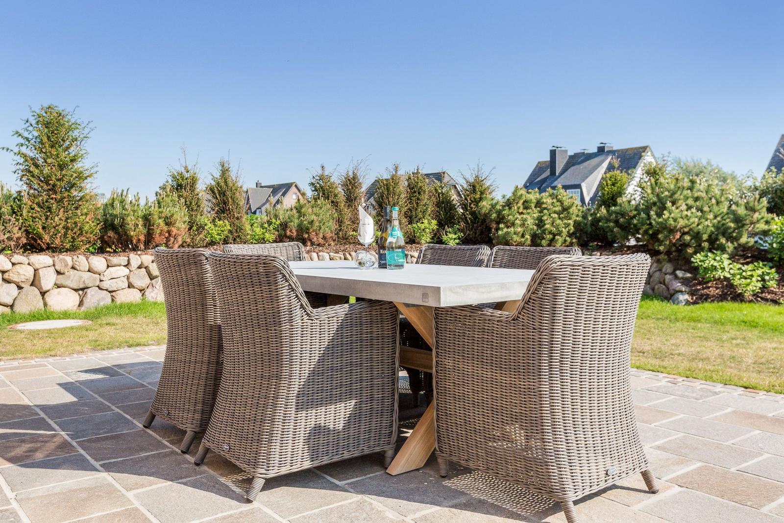 Terrasse mit Esstisch und sechs Stühlen. Hintergrund: Garten, Steinmauer und Häuser.