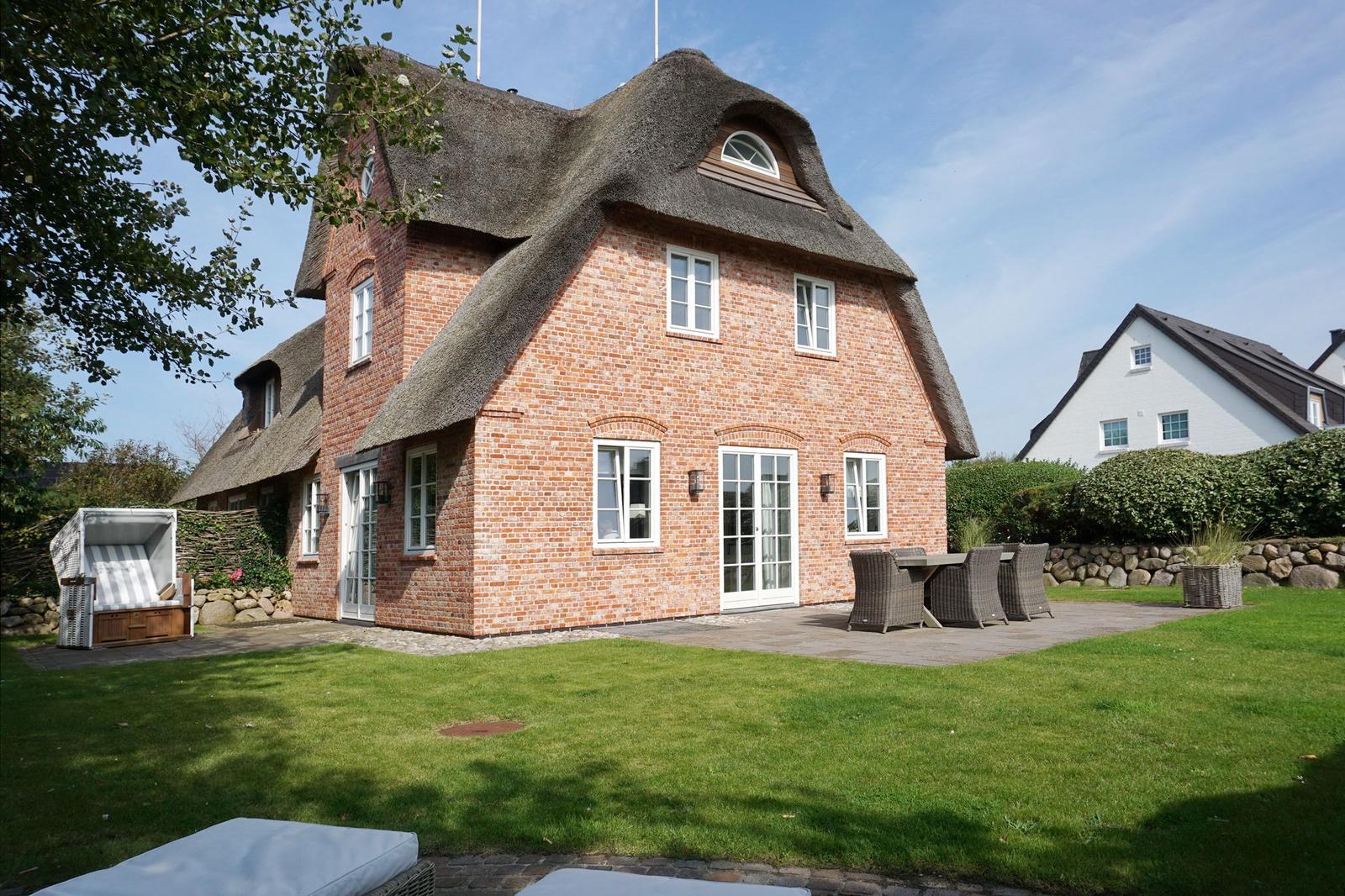 Rotes Backsteinhaus mit Strohdach, Terrasse und Garten mit Liegestühlen.