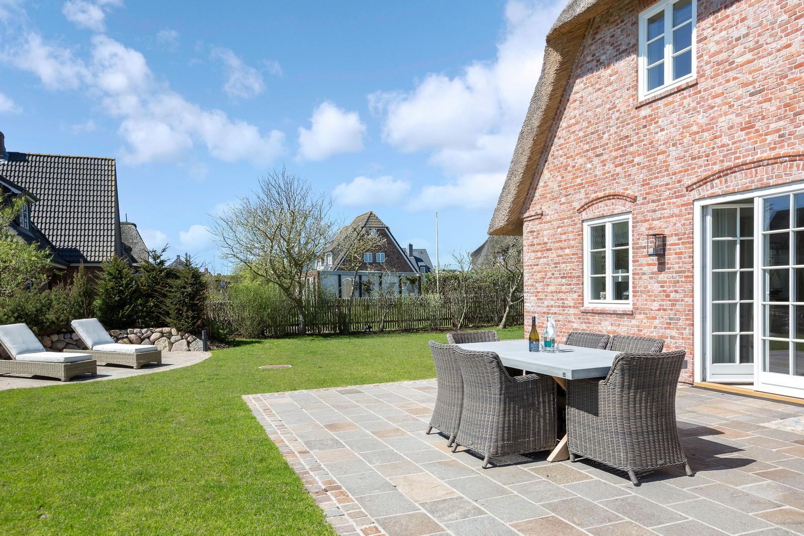 Terrasse mit Tisch und Stühlen neben rotem Backsteinhaus mit Garten und Himmel.
