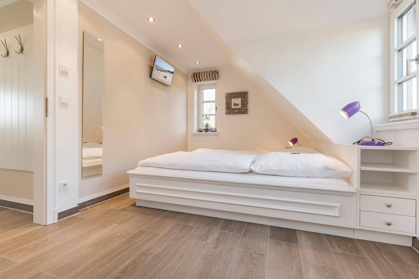 Bedroom with sloped ceiling, white bed, and wooden floor. Windows, mirror, and nightstand with lamp.