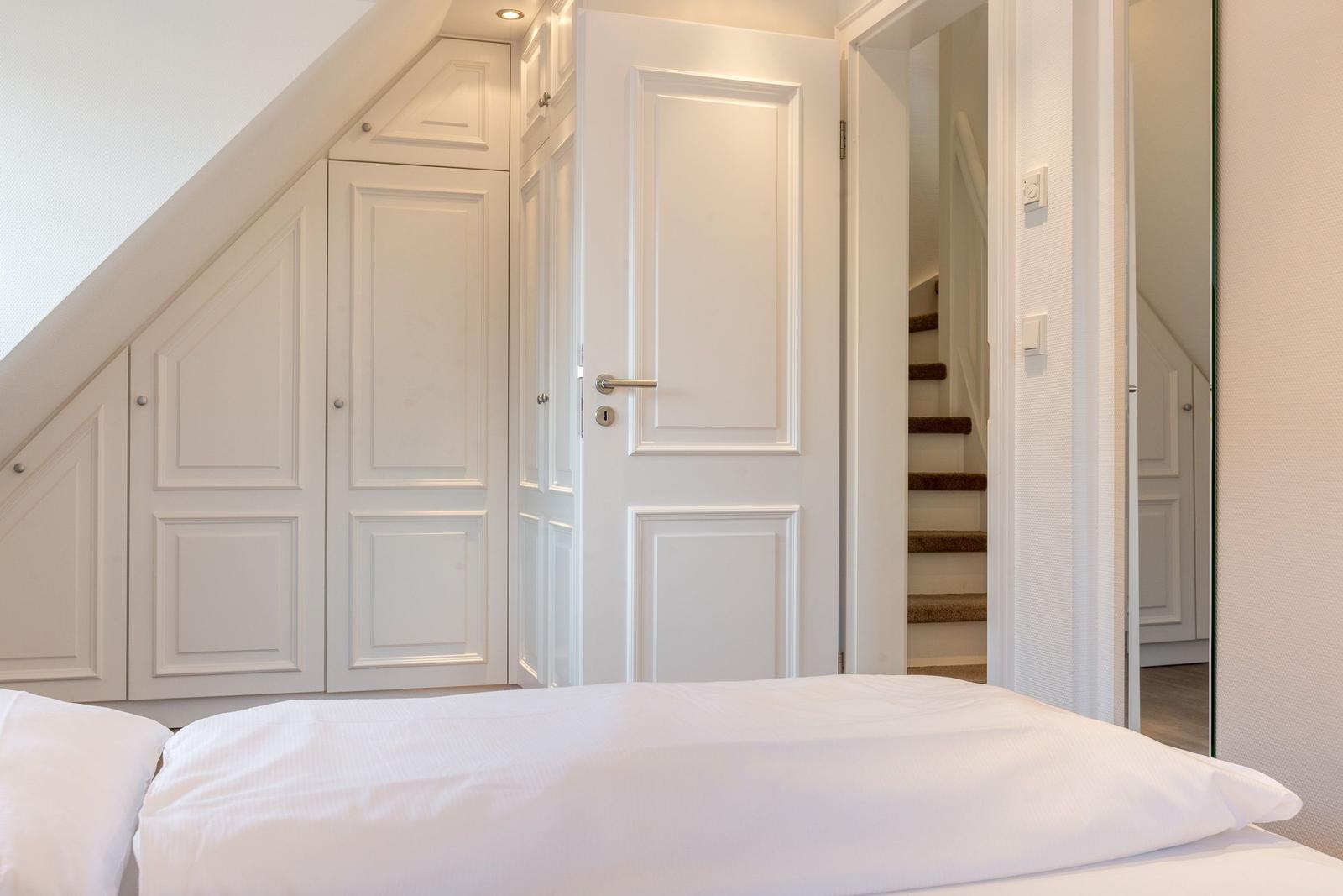 Bedroom with white bed and wardrobe under sloped ceiling. Door leads to stairs.