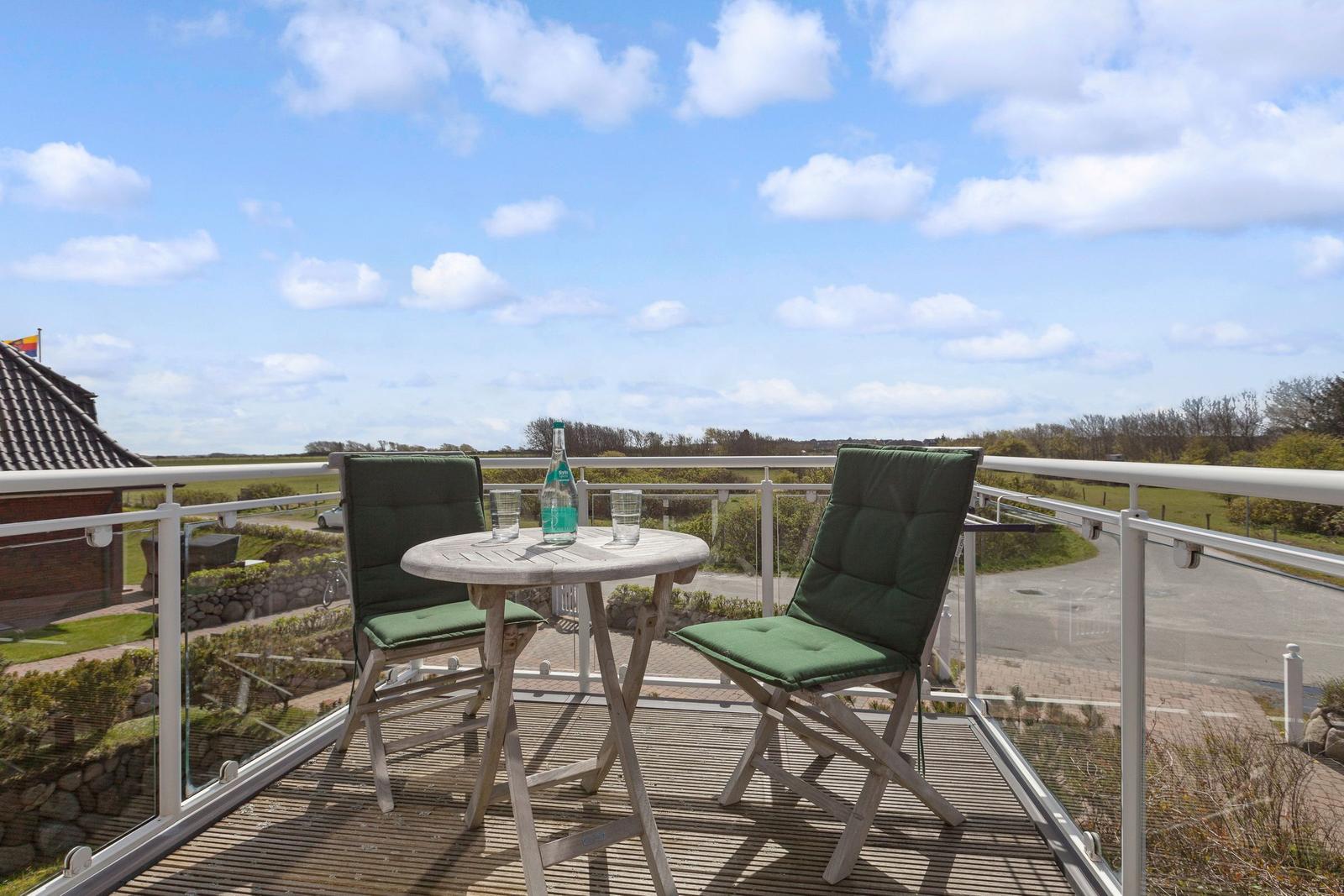 Terrasse mit Tisch und zwei grünen Stühlen. Blick auf Felder und Himmel.