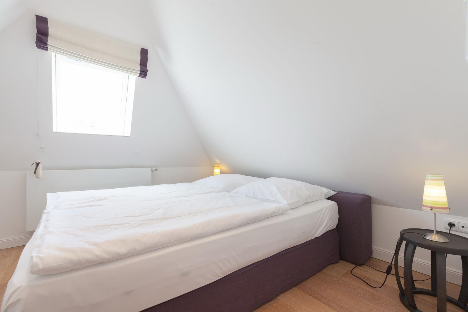 Schlafzimmer mit Doppelbett, Fenster und Nachttisch mit Lampe.