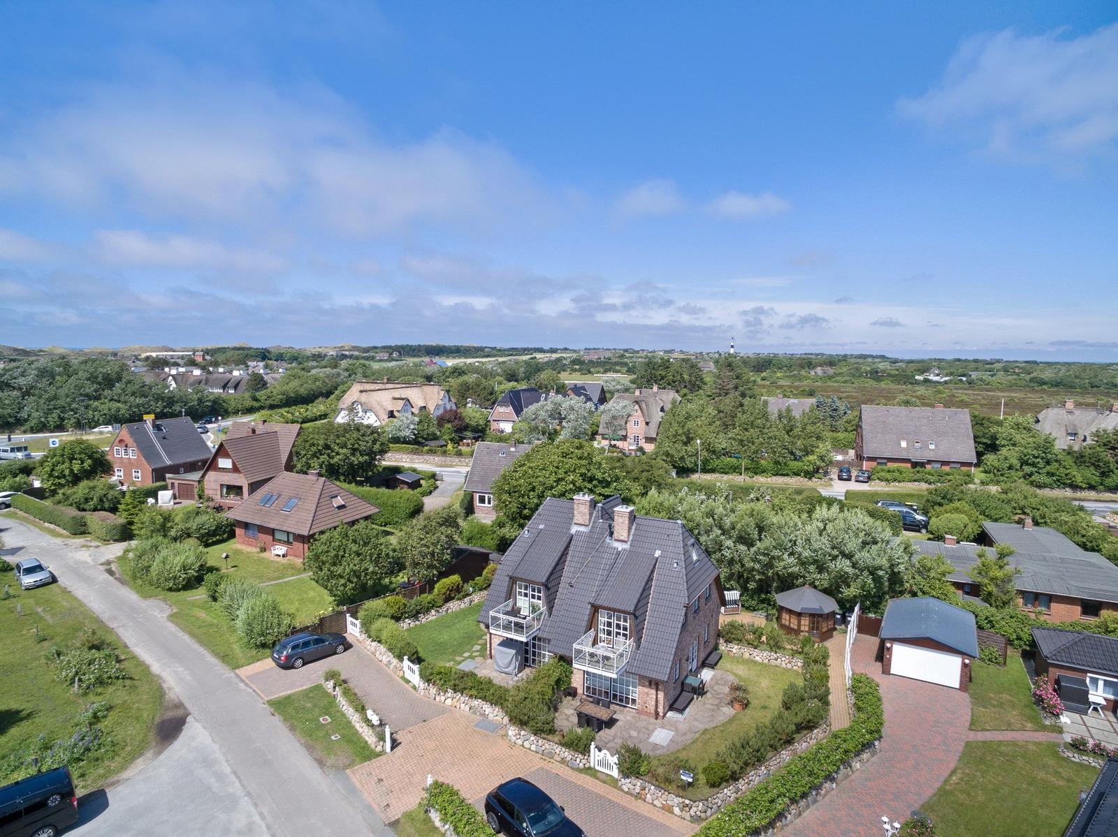 Haus mit Garten und Terrasse, umgeben von Bäumen und weiteren Häusern unter blauem Himmel.